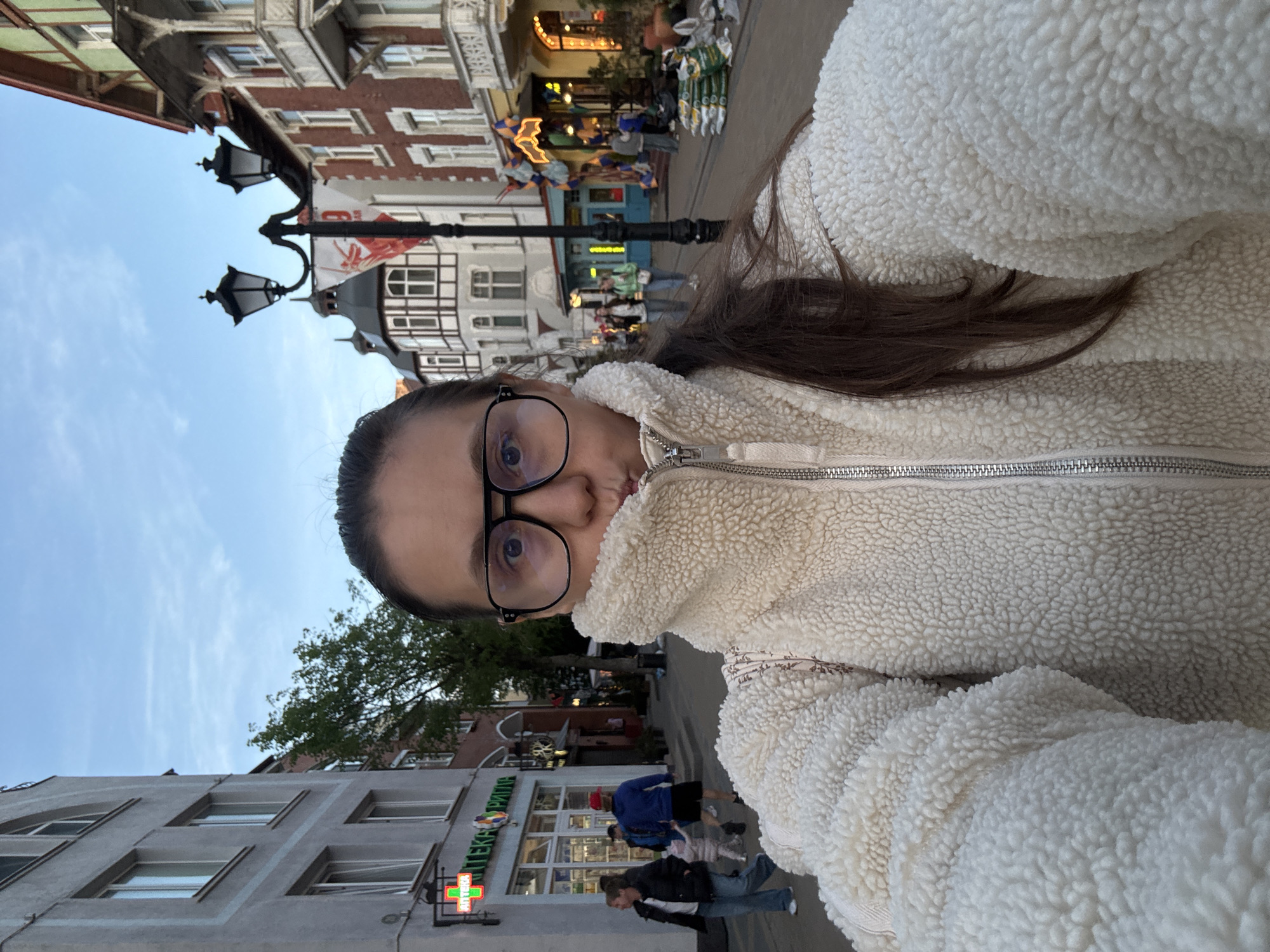 A woman wearing glasses and a white, fluffy jacket takes a selfie. The background reveals a street scene in Zelenogradsk, Russia. A pharmacy is visible in the background. Street lamps and buildings line the street, with other people walking in the background. The sky is partly cloudy.