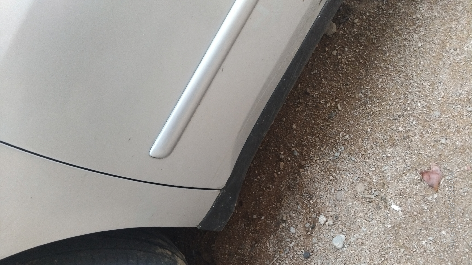 The image shows the lower part of a silver car door and fender, partially obscuring a black plastic trim piece along the bottom edge of the car. The car is parked on a gravel and dirt surface. The background is out of focus, but appears to be a dry, dusty outdoor environment. The scene is located in Howayi, Nigeria. No people are visible in the image.