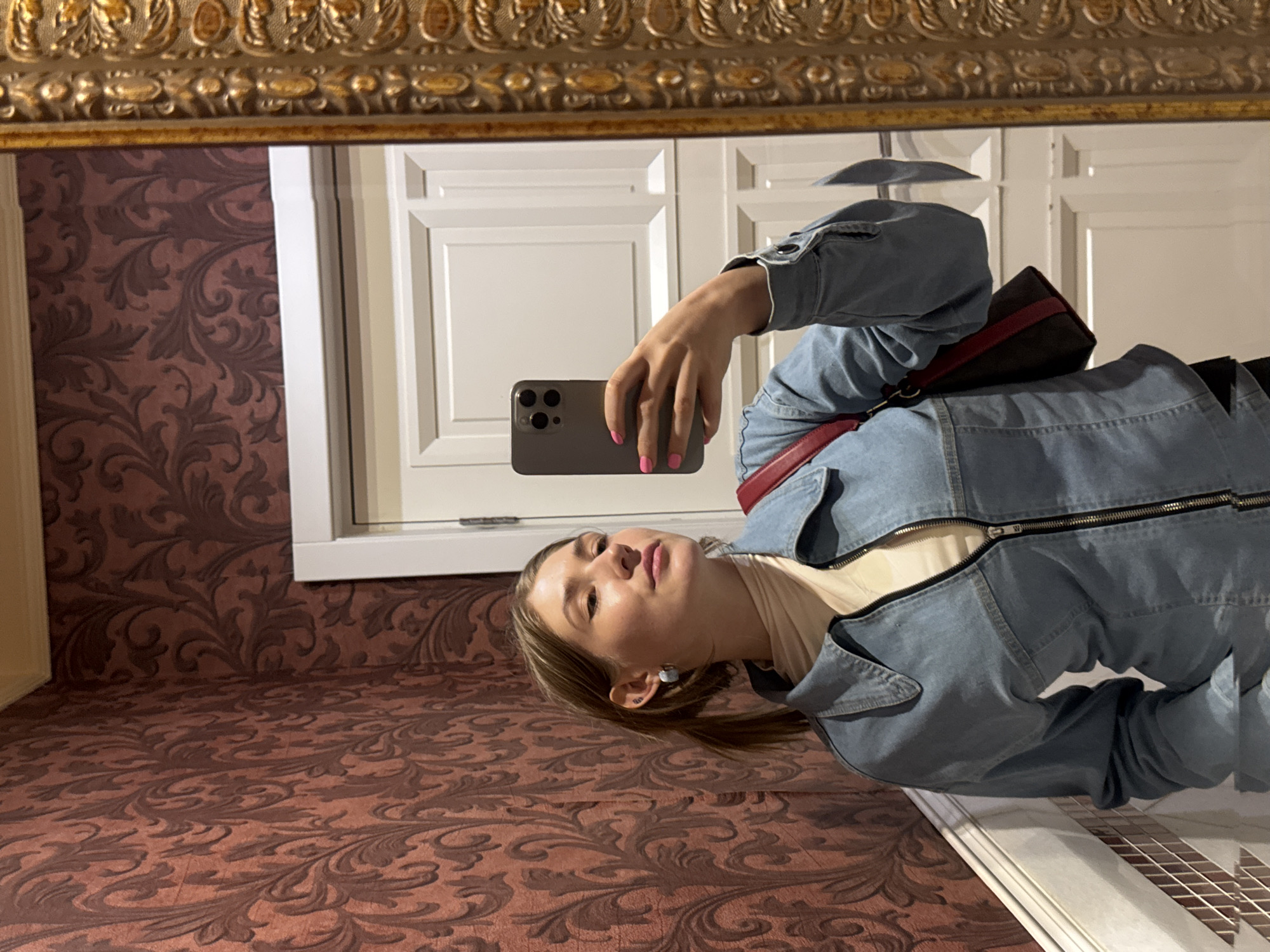 A young woman with blonde hair pulled back in a ponytail, wearing a denim jacket over a cream-colored turtleneck and a crossbody bag, takes a selfie in a mirror. She is in a room with red floral wallpaper and a white door behind her. The mirror has an ornate gold frame. The location is identified as Shkotovo, Russia.