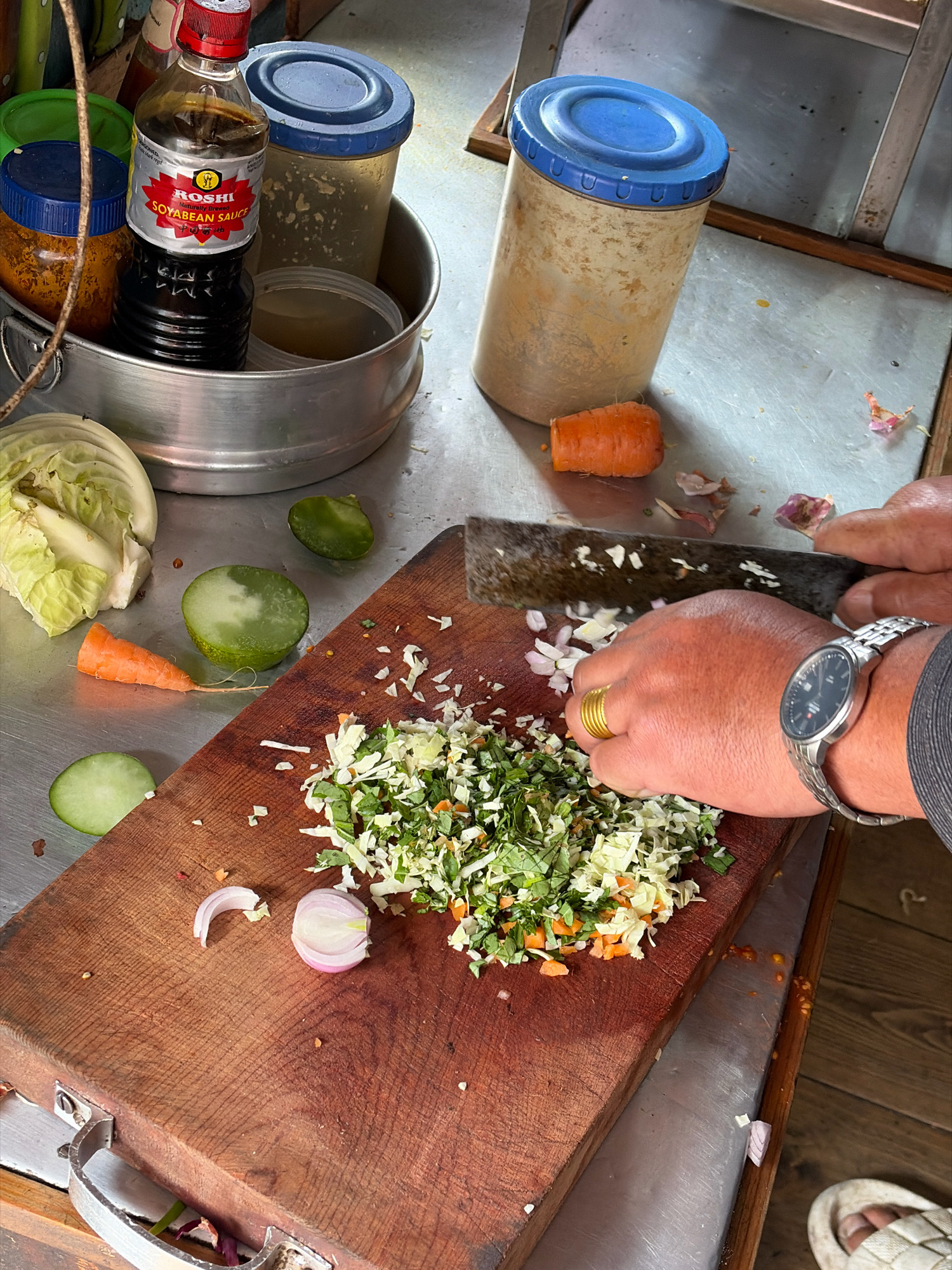 A close-up, overhead shot captures a food preparation scene on a metal surface in Kurung, Nepal. A wooden cutting board is prominently featured, laden with finely chopped vegetables including cabbage, carrots, and what appears to be cilantro. A person's hands are actively chopping more ingredients, likely onions, with a large cleaver. On the left side of the board, whole and sliced vegetables like cabbage, carrots, and cucumbers are visible. To the left of the cutting board, a metal bowl contains a bottle of "ROSHI SOYABEAN SAUCE" and other containers. In the background, two large cylindrical containers with blue lids are present. The scene suggests a domestic or small-scale culinary setting.