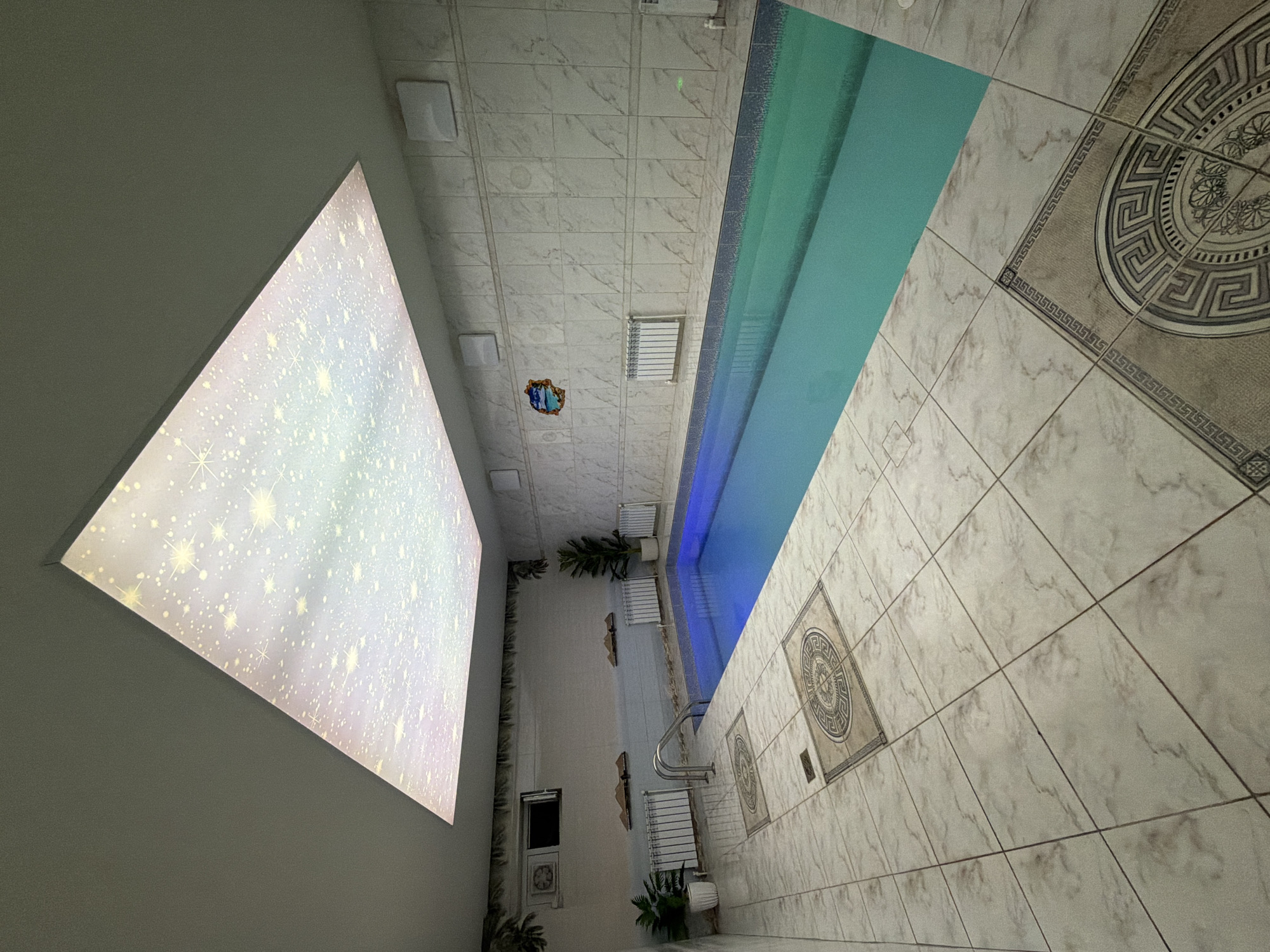 This image depicts an indoor swimming pool facility located in the city of Chaikovsky, Russia. The scene presents a well-maintained, rectangular swimming pool with teal-colored water, featuring an illuminated blue glow at the shallow end where silver metal handrails provide access. The pool's edge is detailed with a darker blue mosaic tile trim. The floor and walls of the room are uniformly covered in light-toned, possibly marble-patterned, ceramic tiles. The floor incorporates decorative square accent tiles with a circular Greek key pattern. The ceiling features a large, rectangular light panel designed to resemble a starry night sky with multi-colored light specks, set within an otherwise light grey ceiling.
The left wall contains a small window, an air conditioning unit, two white radiators, and two potted green plants. Decorative elements resembling foliage are along the top of this wall, and two small, landscape-like wall decorations are mounted below them. The right wall is also tiled and features several square white ventilation grates near the ceiling, one white radiator, and a wall-mounted decorative plaque depicting a small island scene. No people are visible in the image.