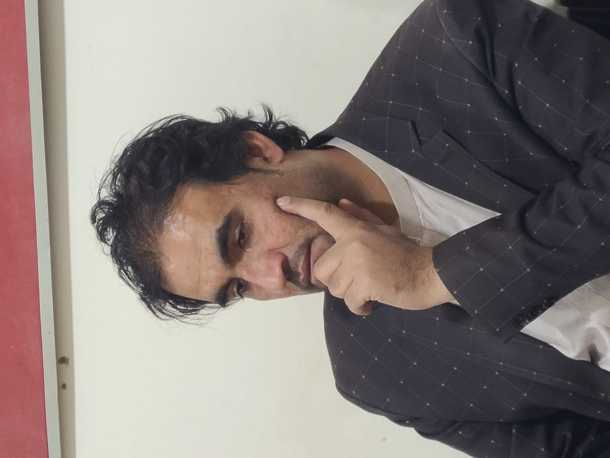 A man is shown from the chest up, in what appears to be an indoor setting with a wall. The man has dark hair and a dark, patterned suit jacket over a white shirt. He is touching his cheek with his finger. The environment includes a white wall and a red object on the left side. There is no information to discern the city or country from the image.