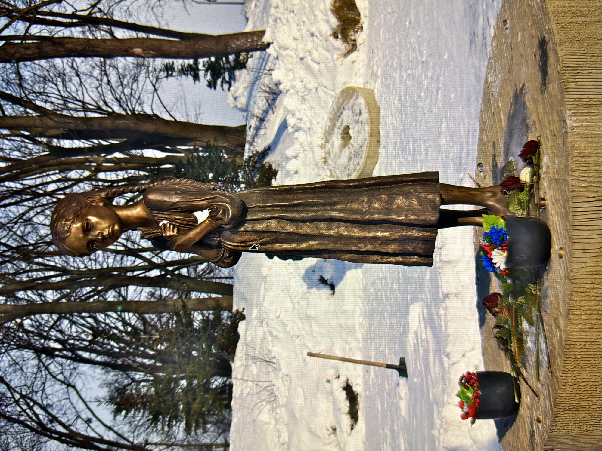 The image shows a bronze statue of a young girl holding a sheaf of wheat, standing on a circular stone pedestal covered in snow. The girl is dressed in a long, dark robe, and her hair is braided. She is standing in an outdoor setting with snow-covered ground and bare trees in the background. There are two black pots filled with red and blue flowers, and several red roses and greenery placed on the pedestal around the base of the statue. A shovel with a wooden handle is stuck in the snow to the left of the statue. A stone millstone is visible in the snow to the right of the statue. This is the "Girl with the Ears of Wheat" monument, located in Kyiv, Ukraine.