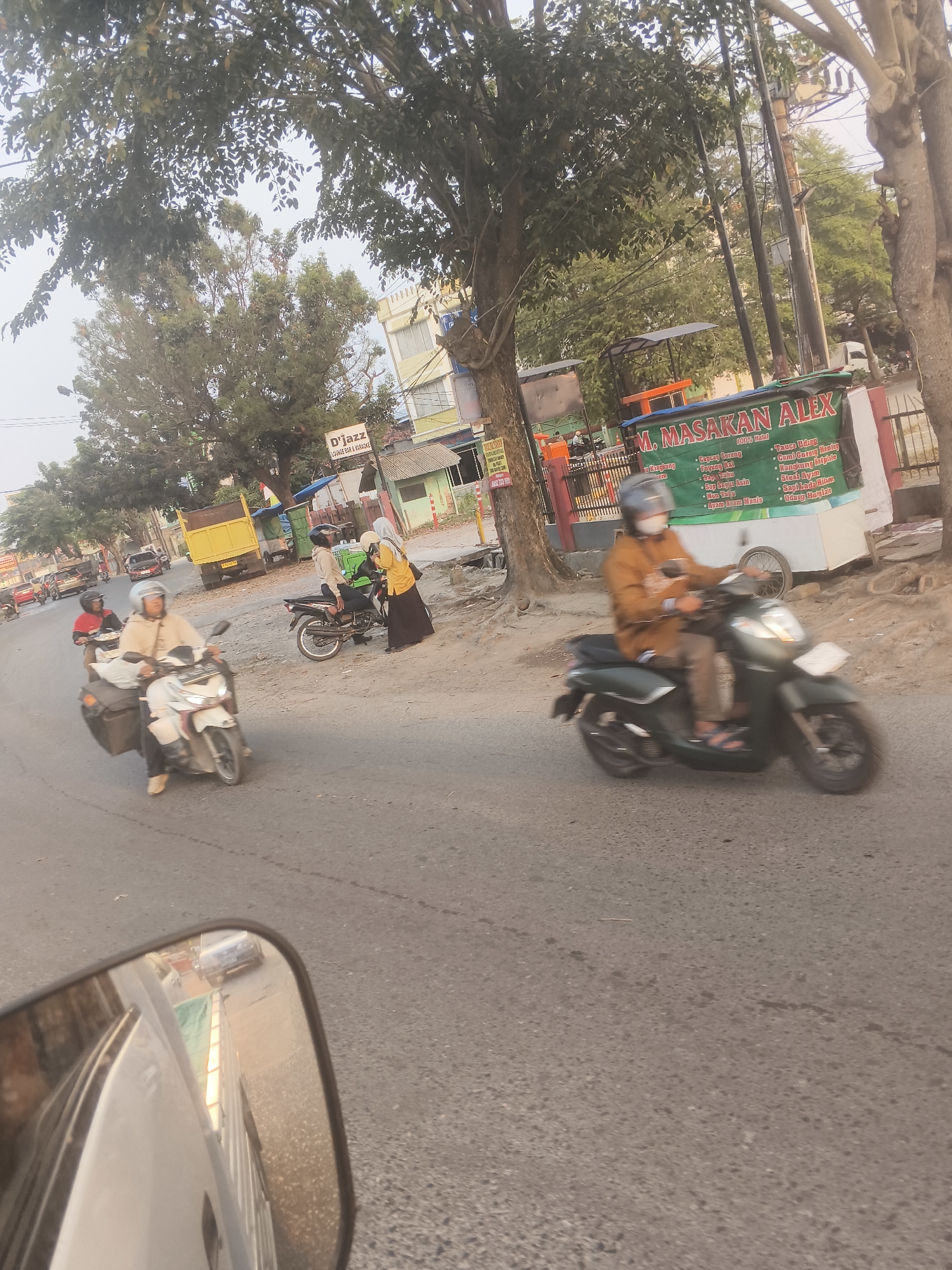 The image depicts a street scene in Bandar Lampung, Indonesia, likely in the morning or late afternoon given the lighting. The foreground shows a portion of a white vehicle's side mirror reflecting the road and the side of the vehicle itself. The road is paved and appears to be a two-lane street with moderate traffic.
Several people are present, mostly on motorcycles. On the left side of the road, a person wearing a white helmet and a beige jacket is riding a white scooter. Behind them, another person in a red shirt and helmet is riding a motorcycle. Further up the road on the left, a yellow truck is parked.
In the middle ground, a woman in a yellow hijab and black skirt is standing near a parked motorcycle, interacting with another person who is also on a motorcycle. The person on the motorcycle is wearing a light-colored jacket and a white helmet.
On the right side of the road, a person wearing a brown jacket and a blue helmet is riding a dark green scooter. Behind them, there's a food stall with a sign that reads "M. MASAKAN ALEX" and lists various food items. Adjacent to the food stall is a building with a sign that says "D'Jazz LOUNGE BAR & KARAOKE."
The environment is characterized by trees lining the street, power lines, and various buildings, suggesting an urban or semi-urban setting. The overall atmosphere is that of a typical street scene with local commerce and transportation.
