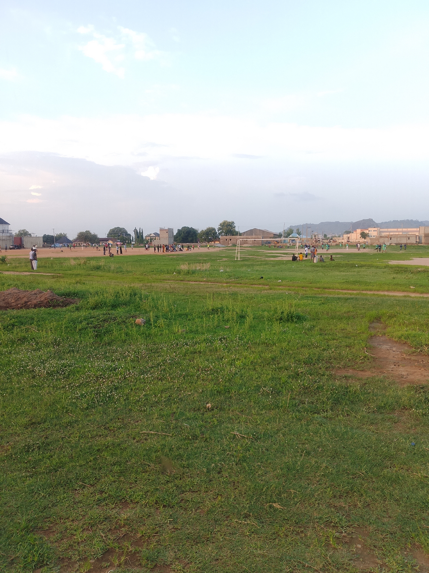 The scene depicts a grassy field in Ibo, Nigeria. In the foreground, the grass is green and somewhat overgrown, with patches of bare earth visible in the lower right. In the mid-ground, people are engaged in various activities, including what appears to be a volleyball game and other undefined sporting actions. Several buildings and structures are visible on the horizon, including a stone tower and some residential or commercial buildings. A hill is present in the far background. The sky is overcast.