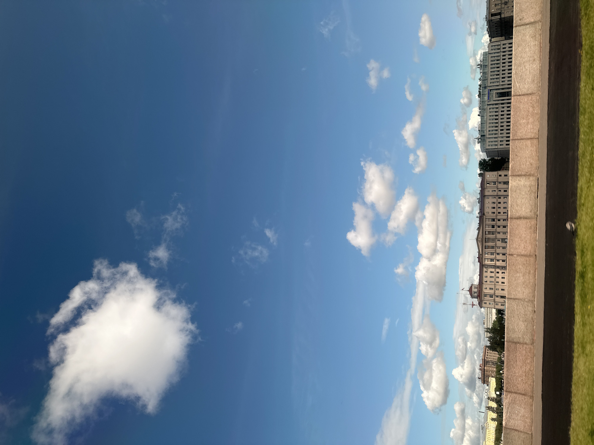 The image captures a wide-angle view of the Neva River embankment in Saint Petersburg, Russia, on a clear day with scattered clouds. In the foreground, a concrete barrier separates the embankment from a grassy area and a paved surface, possibly a road. Across the river, a line of grand, historic buildings forms the city's skyline. These buildings are characterized by their classical architecture, with multiple stories, symmetrical windows, and ornate details. Some buildings feature domes or towers, indicative of their official or historical significance. The sky above is a vibrant blue, with a few fluffy white clouds, one particularly large cumulus cloud dominating the upper left portion of the frame. The overall impression is one of a typical sunny day in Saint Petersburg, showcasing its majestic architecture and the expansive sky over the Neva River. There are no people or animals clearly visible in the image.