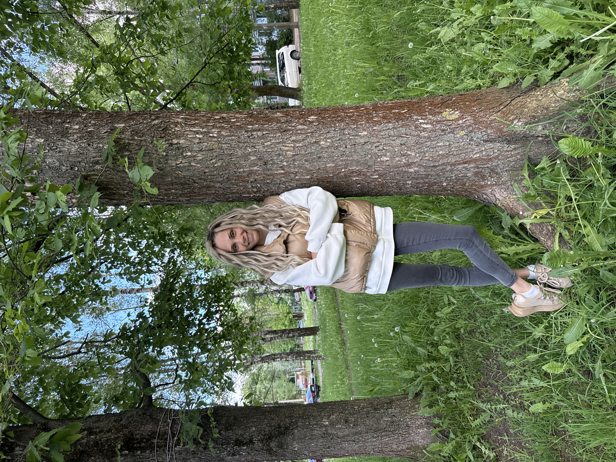 A young woman with long blonde dreadlocks is leaning against a tree trunk in a grassy area. She is wearing a white hooded sweatshirt, a beige puffy vest, grey jeans, and tan sneakers. She is smiling at the camera. The location appears to be a park or green space, with trees visible in the background. There are buildings and parked cars in the distance, possibly indicating an urban or suburban environment.
Based on the provided information, it is in the city of Rusanovo, Russia.