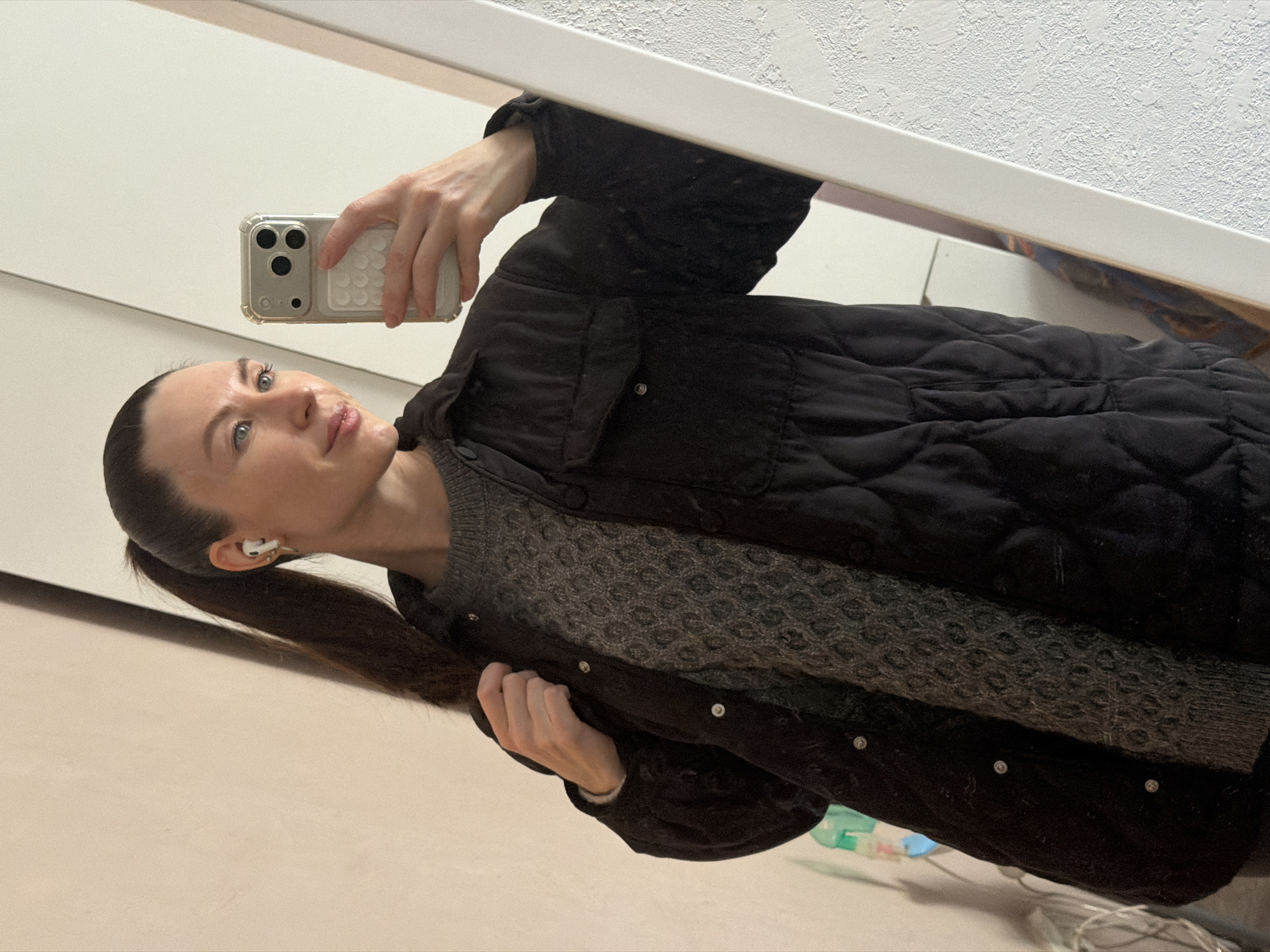 A woman is captured in a mirror selfie, reflected from a slightly tilted angle, giving the impression of her lying horizontally. She has dark hair pulled back into a ponytail and is looking subtly upwards and to the side with a calm expression and a faint smile. She is wearing a dark, possibly black, quilted jacket over a grey sweater with a textured, patterned knit. A white wireless earbud is visible in her right ear.
She holds a silver or light-colored smartphone with three camera lenses (likely an iPhone Pro model) in her right hand, encased in a clear cover with a textured white grip or accessory on the back. The background reflected in the mirror consists primarily of plain light-colored walls, one with a subtle textured finish, and a narrow white horizontal element, possibly a shelf or door frame. Towards the bottom right, some indistinct items and cables are visible on what appears to be a surface or floor. The lighting suggests an indoor setting, likely artificial. There is no visible text in the image. The location context provided is Perm, Russia.