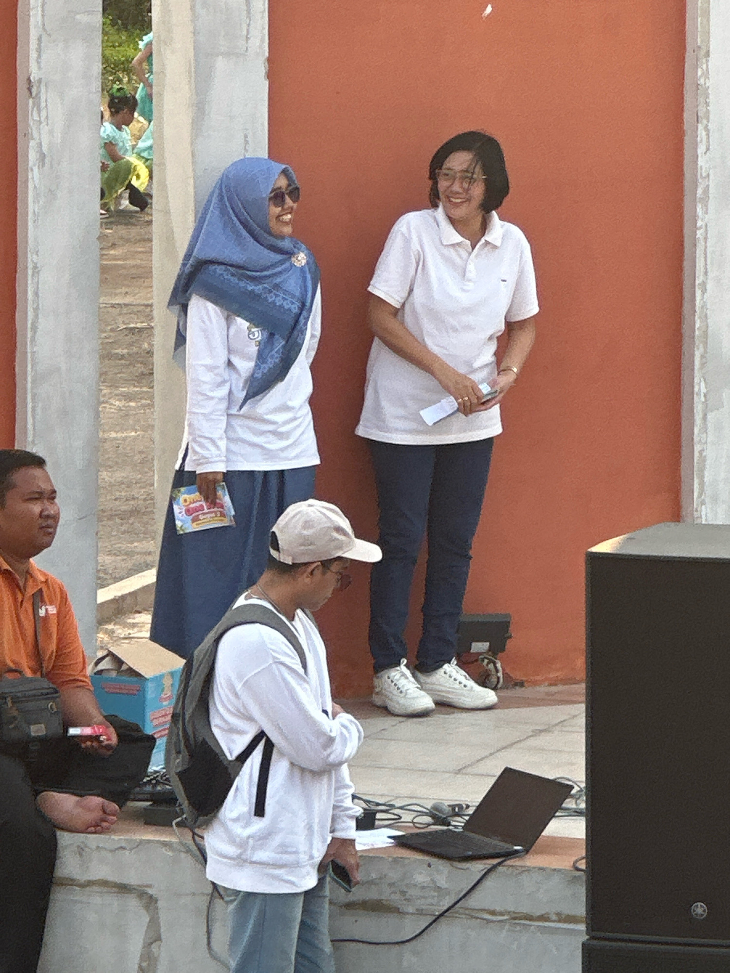 In Ngingasrembyong, Indonesia, a medium shot captures an outdoor scene with three individuals and a large black speaker. To the left, a man wearing an orange polo shirt sits with his left foot bare. Next to him, a woman in a blue hijab and sunglasses smiles, holding a small booklet. To her right, another woman in a white polo shirt and dark blue pants smiles, holding a piece of paper and a pen. In the foreground, a person wearing a white long-sleeved shirt and a light-colored baseball cap faces away from the camera, with a backpack on their shoulders and a laptop on a ledge in front of them. The background features an orange wall, a concrete structure, and blurred figures of people in the distance.