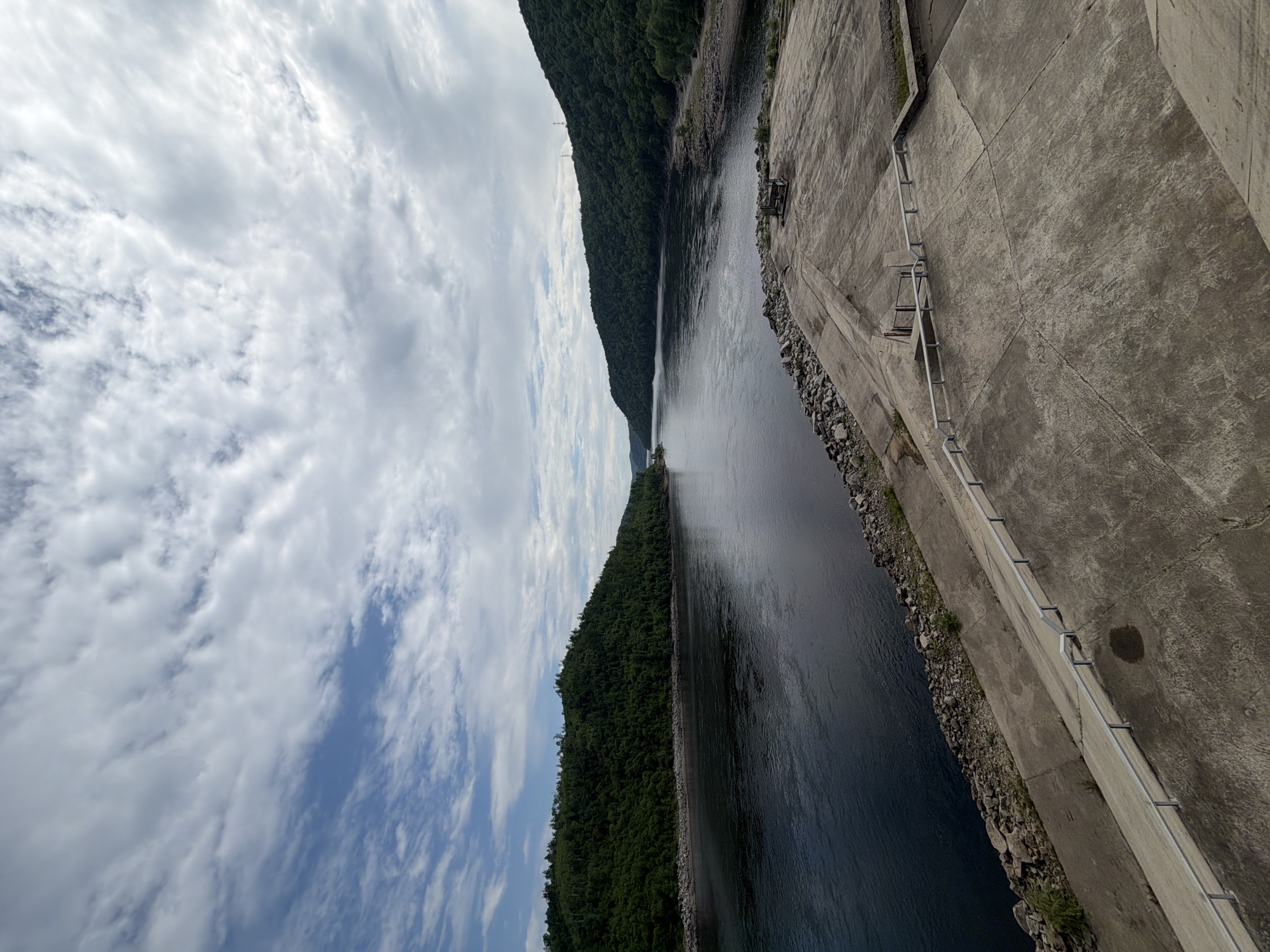 A wide-angle, vertical shot captures a serene landscape under a partially cloudy sky. The scene features a large body of dark water, likely a river or reservoir, bordered by densely forested hills. In the foreground, a concrete structure with a gradual incline leads down towards the water's edge. This concrete area is partially covered with scattered rocks and features a metal railing along its perimeter. A small wooden bench is visible on the concrete platform. The water is calm, reflecting the sky and surrounding greenery. The overall impression is one of natural beauty and tranquility, potentially a scenic overlook or dam area. There are no people visible in the image. The location is identified as the Talakan reservoir in Talakan, Russia.