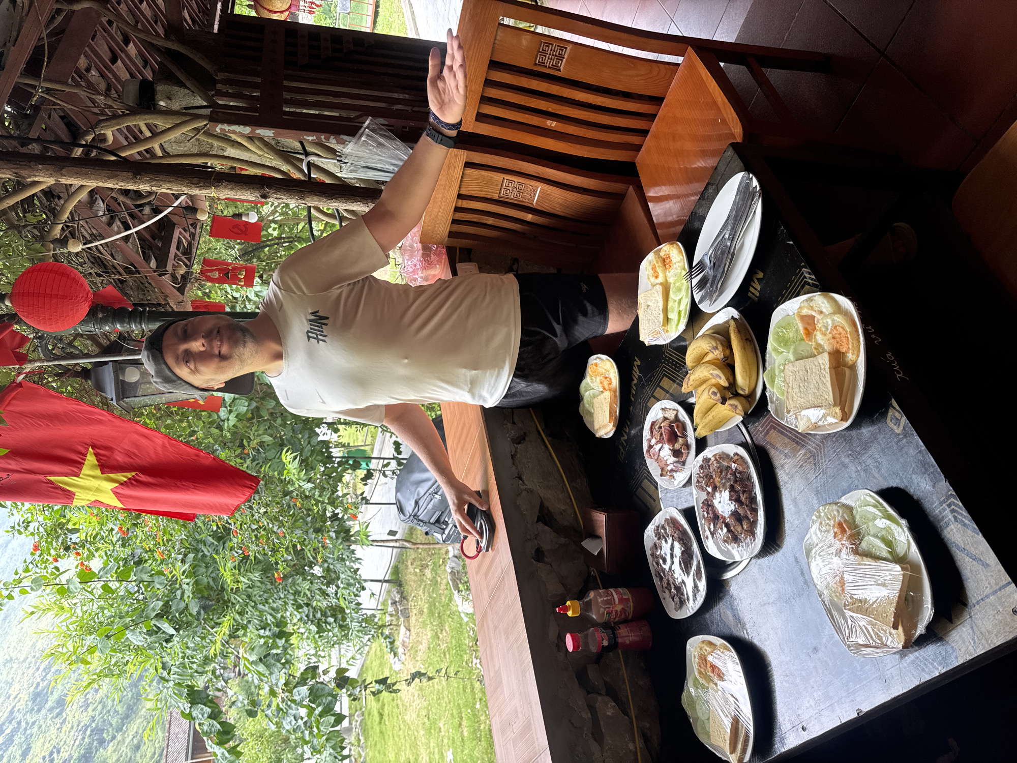 The image captures a scene in Mèo Vạc Commune, Vietnam. A man stands beside a wooden table laden with various dishes, some covered in plastic wrap. The meals include sandwiches, fruit, and meats. Utensils are also visible. He wears a light-colored shirt, dark shorts, and a cap and holds his hand up. A Vietnamese flag hangs near a dense green plant with red flowers. In the background, there is a glimpse of the surrounding landscape and structures, including wooden furniture. The setting seems to be an outdoor covered area.
