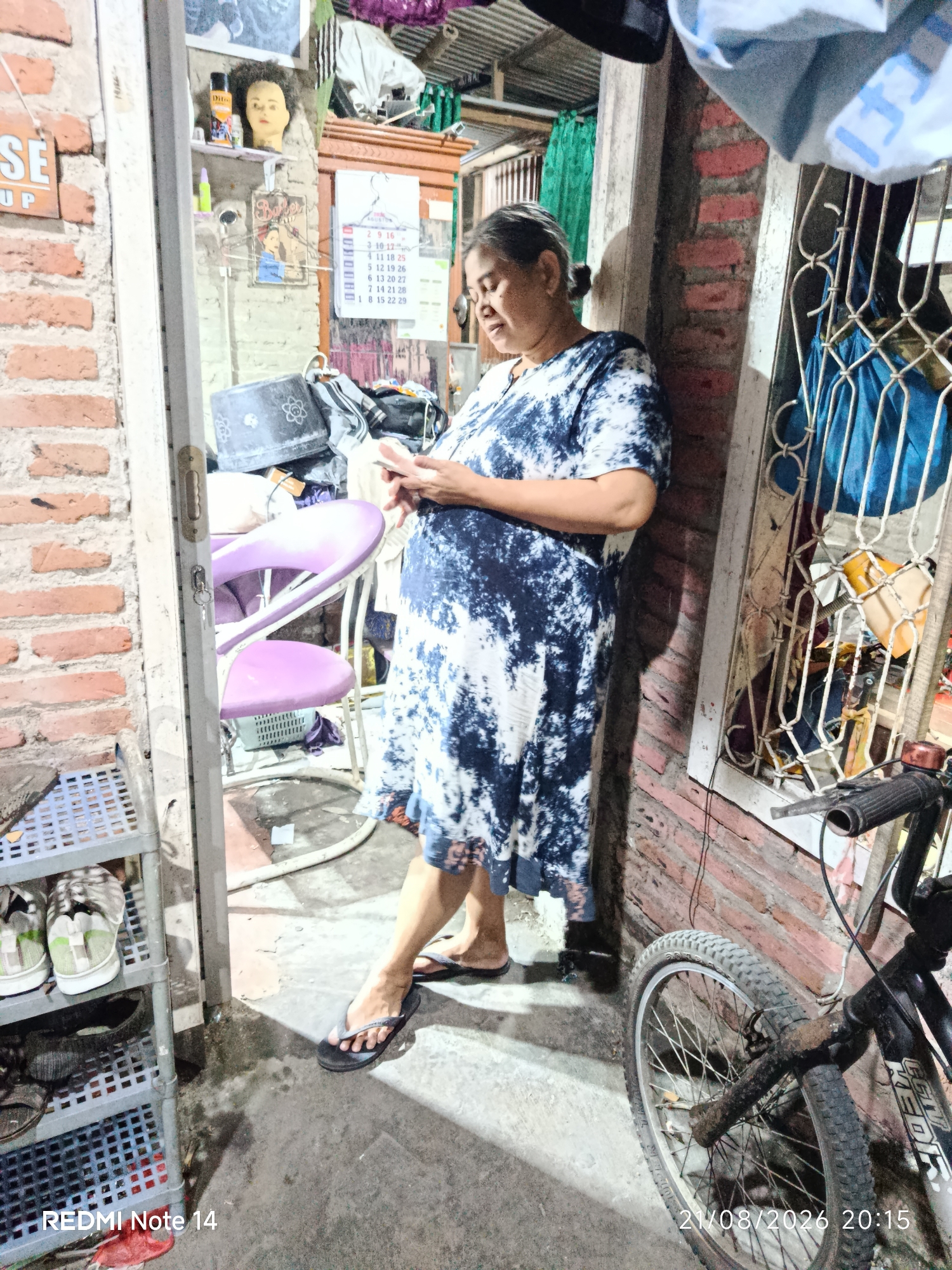 A woman with dark hair pulled back is standing in a doorway, leaning against a brick wall. She is wearing a blue and white tie-dye dress and dark flip-flops. She is holding a mobile phone in her hands and looking down at it. Next to her, a bicycle with a black frame and visible rust is parked. The doorway opens into a cluttered indoor space with shelves holding various items, including shoes, and purple chairs are visible behind the woman. A calendar with handwritten dates is on the wall. Outside the doorway, a grey plastic shelving unit holds two pairs of shoes. The ground is concrete and appears worn. The timestamp in the lower right corner indicates the image was taken on 21/08/2026 at 20:15.