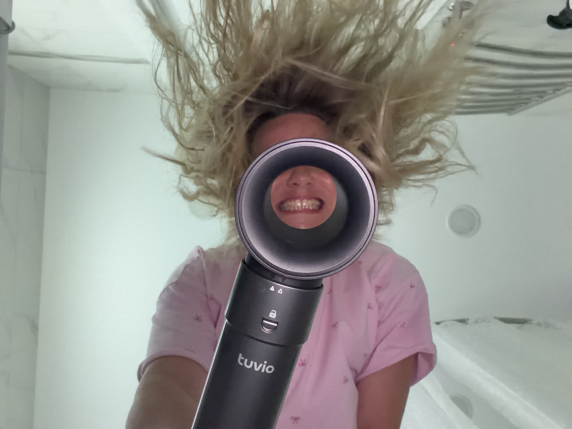 An upward-angled, close-up photograph depicts a person, likely a woman, with voluminous, light blonde hair standing dramatically on end, as if blown by strong air. The subject, wearing a light pink, short-sleeved top with faint patterns, holds a dark grey "tuvio" brand hair dryer pointed directly towards the camera. Through the hair dryer's circular nozzle, the person's mouth and visible teeth are seen in a broad, cheerful smile, suggesting a moment of playful amusement. The handle of the hair dryer clearly displays the "tuvio" logo, accompanied by symbols including two upward-pointing triangles and a padlock icon. The background reveals a bright indoor setting, characteristic of a bathroom in Votkinsk, Russia, featuring white tiled walls, a ceiling light, and what appears to be a metallic towel warmer on the right. The image captures a humorous and energetic interaction with the hair dryer.