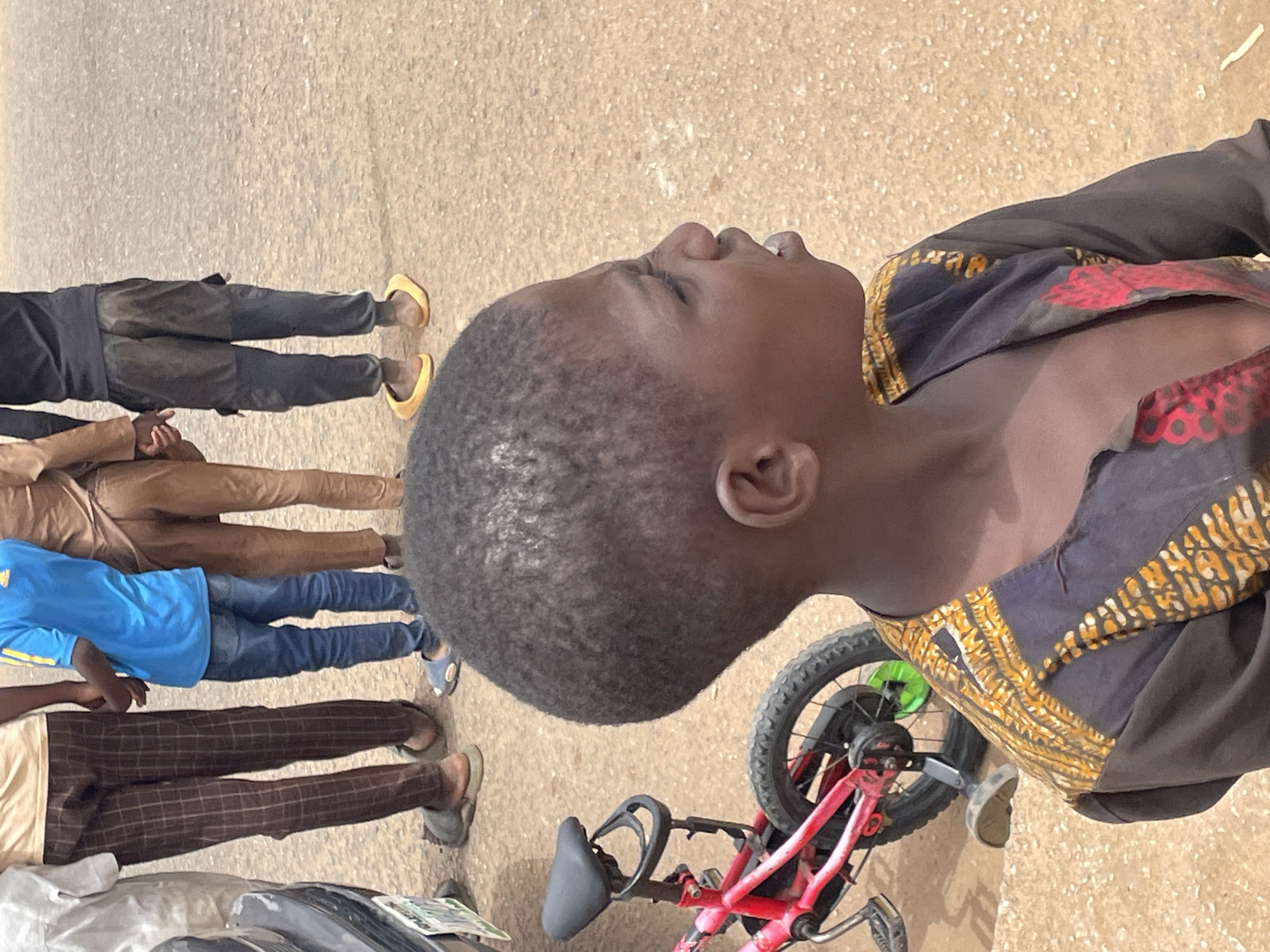 The image shows a group of people standing on a paved surface, likely a street. A child is in the foreground, wearing a patterned shirt and standing near a red bicycle. Behind the child, other people stand in a line. They are wearing various clothing styles, including casual pants, jeans, and sandals. The environment appears to be outdoors in daylight. The location is Jos, Nigeria.