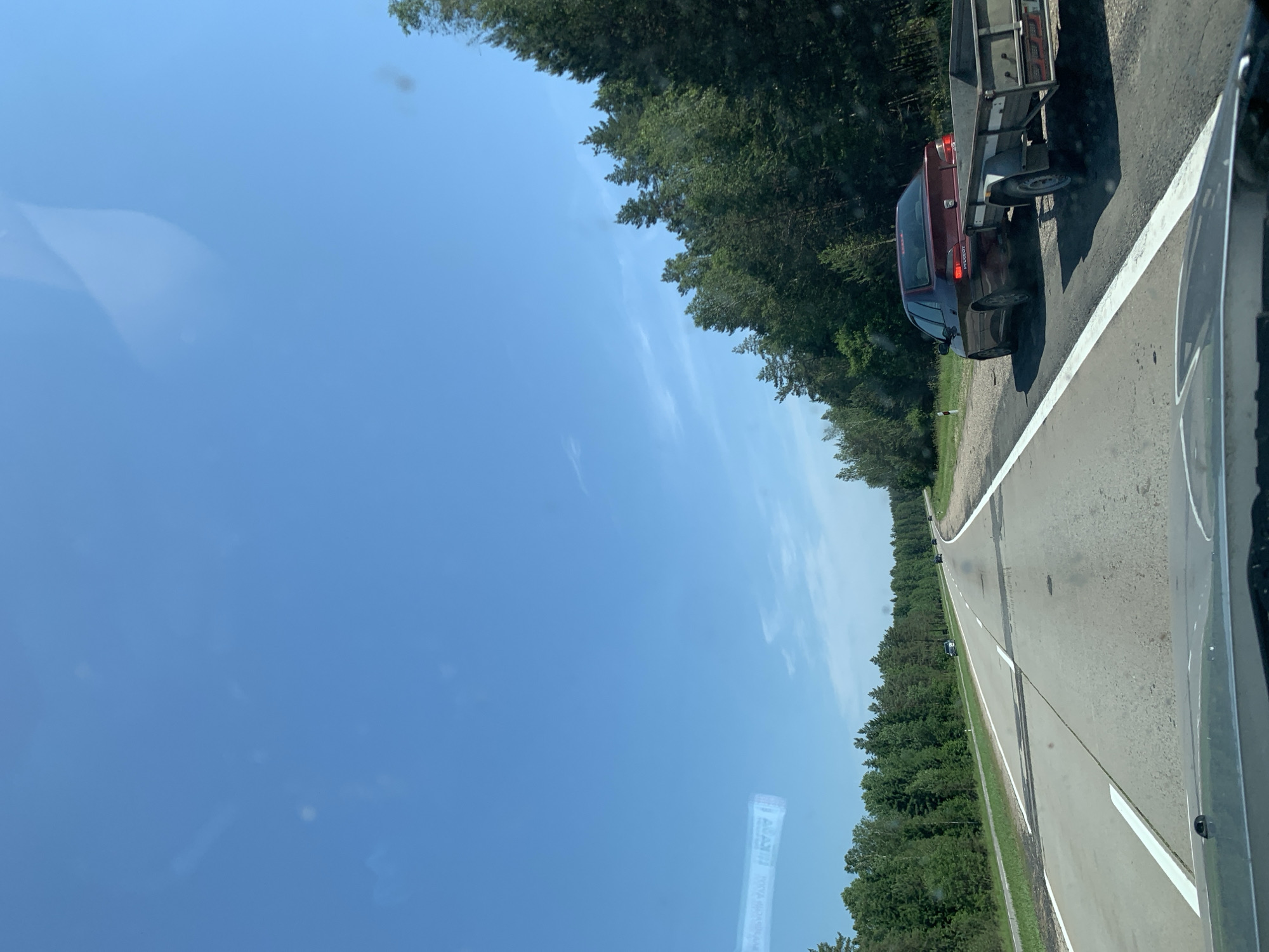 The image shows a view from inside a vehicle traveling on a paved two-lane road, likely a highway, with white lane markings. The road curves gently to the right, bordered by a grassy shoulder and a dense forest of green trees on both sides. The sky is clear blue with a few wispy clouds. On the right side of the road, a dark red Peugeot sedan is pulling a small utility trailer. Further ahead on the road, other vehicles can be seen in the distance. The scene appears to be set in a rural or semi-rural area in Belarus, given the mention of Гацукоўскі сельскі Савет, which is a rural council in the Slutsk District of Minsk Region, Belarus. The weather is bright and sunny.