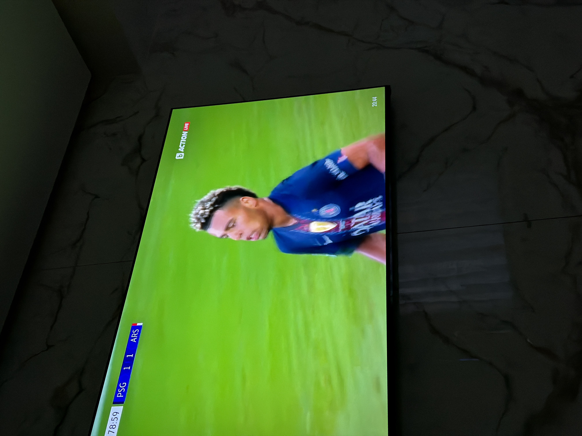 The image shows a television screen displaying a live football match. The score is PSG 1, ARS 1. The time on the screen is 78:59 and 20:44. A soccer player is visible in the center of the screen. The TV is placed near a wall with a marble-like texture. The location of the image is Asenovgrad, Bulgaria, as indicated in the image title.