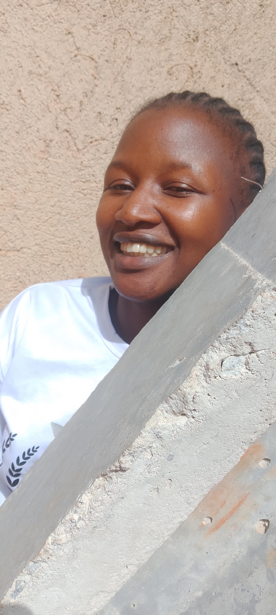 A woman with dark skin and braided hair smiles from behind a concrete support beam in what appears to be an outdoor setting. She wears a white t-shirt with a leafy design. The background is a textured beige wall. Details regarding the city of Jos, Nigeria, cannot be independently confirmed from the image.