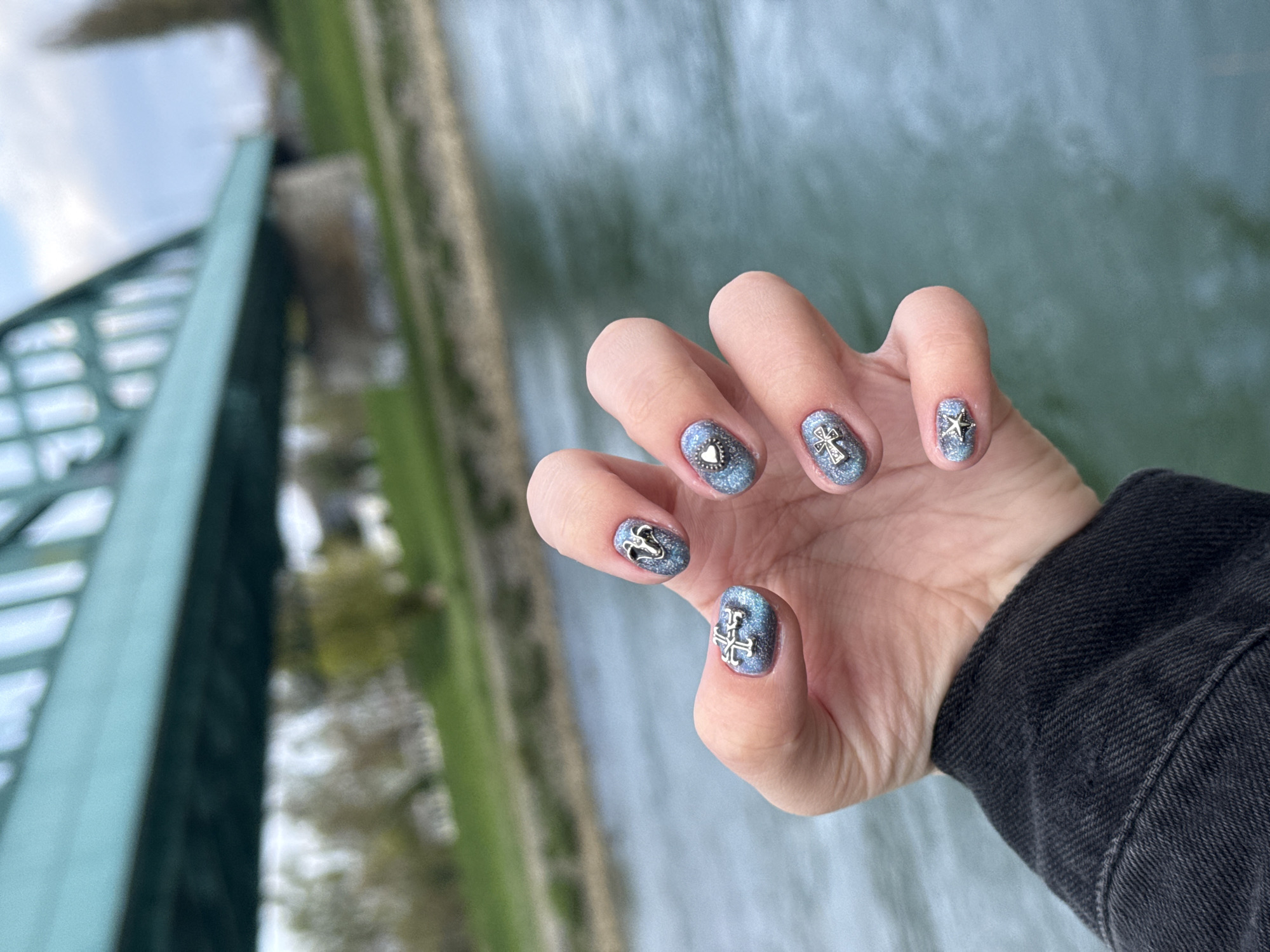The image captures a hand held over a bridge in Zagreb, Croatia. The hand belongs to a person wearing a dark denim jacket, focusing on their unique nail art. Each nail features a shimmery blue base and is decorated with a small, silver charm: a heart, a cross, a star, an anchor, and another cross. The backdrop showcases the Mrežnički Bridge, recognizable by its distinctive green structure and the flowing river below, indicating the city of Zagreb, Croatia. The overall scene gives an impression of a casual, urban setting with a personal touch added through the nail art.