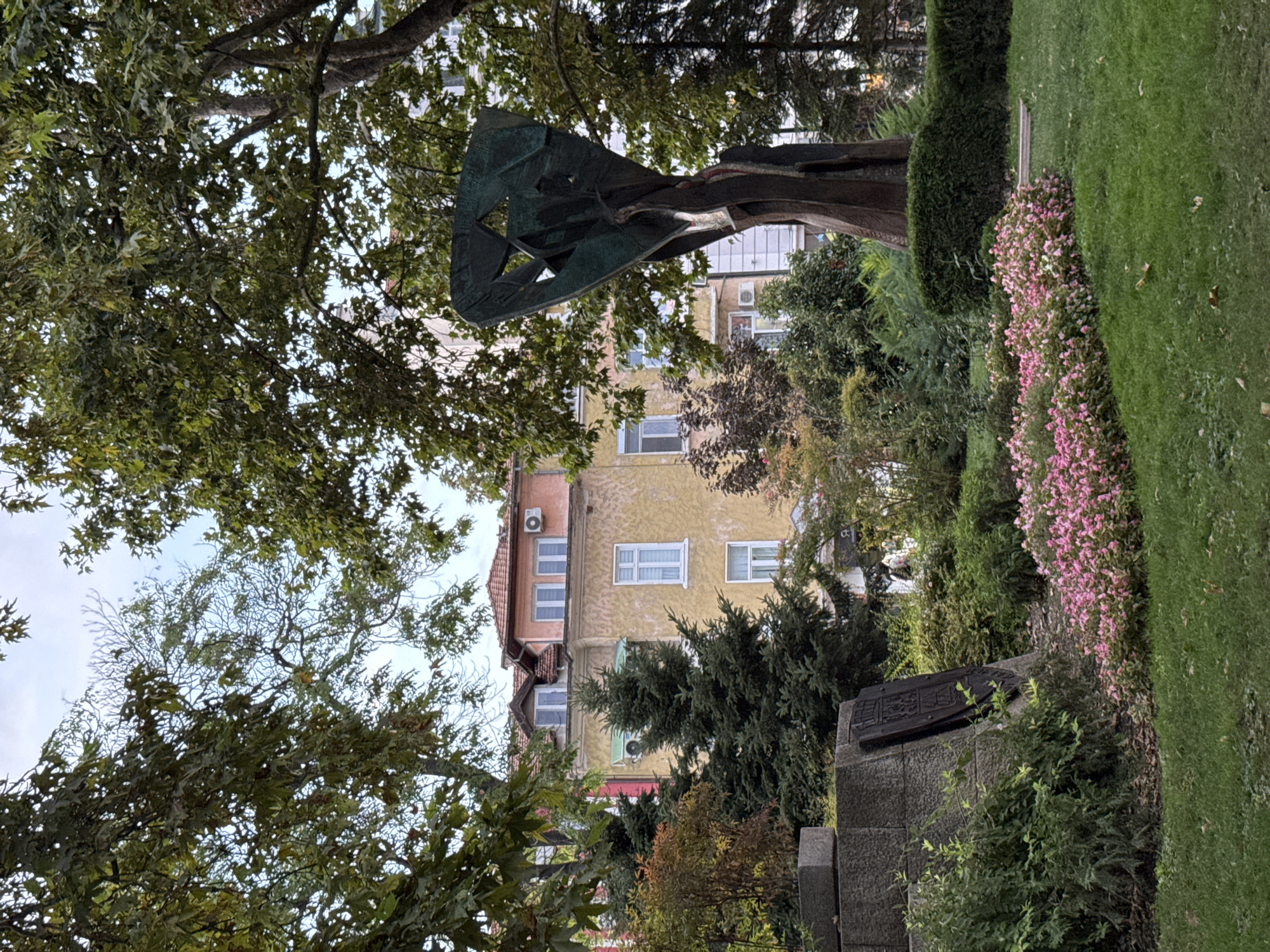 This image depicts a park scene in Plovdiv, Bulgaria, likely near the city center given the architectural style of the buildings. In the foreground, a manicured lawn is bordered by a bed of vibrant pink flowers. To the left, a stone monument with a dark, embossed plaque is partially obscured by lush green bushes. A large, abstract metallic sculpture, appearing to be made of bronze or a similar material with a green patina, stands prominently in the mid-ground. Behind the sculpture and monument, a multi-story building with a light yellow facade and a tiled orange roof is visible, featuring multiple windows and an air conditioning unit. Evergreen trees, including a tall spruce, and deciduous trees with green and autumnal foliage are scattered throughout the scene, framing the view of the building and sculpture. The sky is overcast with a light blue hue. No people are clearly discernible in the image.