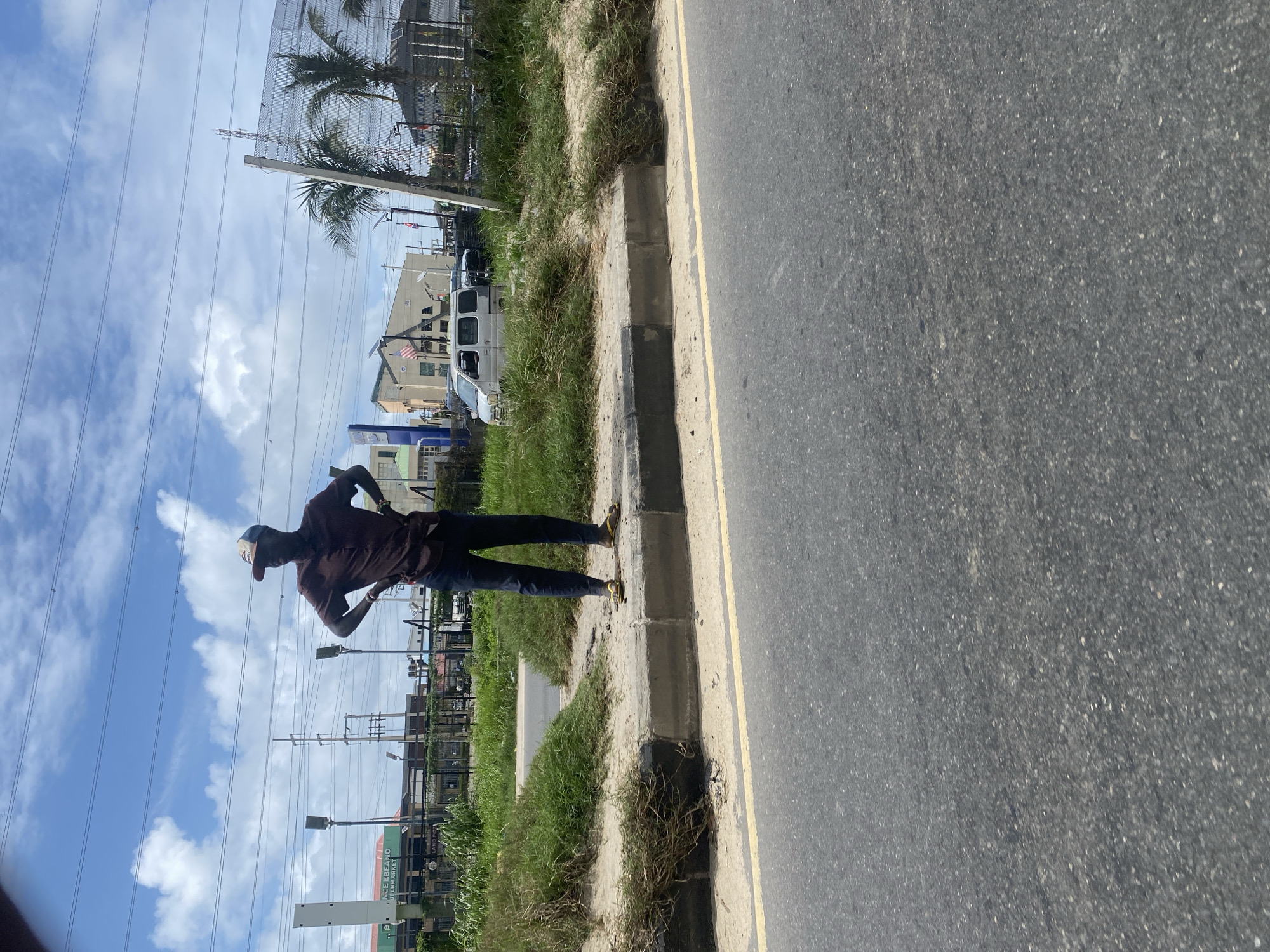 In Lekki Peninsula II, Nigeria, a person stands on the side of a road next to a sidewalk. The individual is wearing a brown shirt, dark pants, and yellow flip-flops, with a cap on their head. Their hands are on their hips, and they are facing forward. Behind them, there is a grassy area with some dry brush, a road, and in the distance, several buildings, including what appears to be a supermarket. Power lines and utility poles are visible in the sky, along with scattered clouds. A white van is parked on the side of the road in the background, and palm trees and a fence are also present. The overall scene depicts a typical street view in an urban environment.