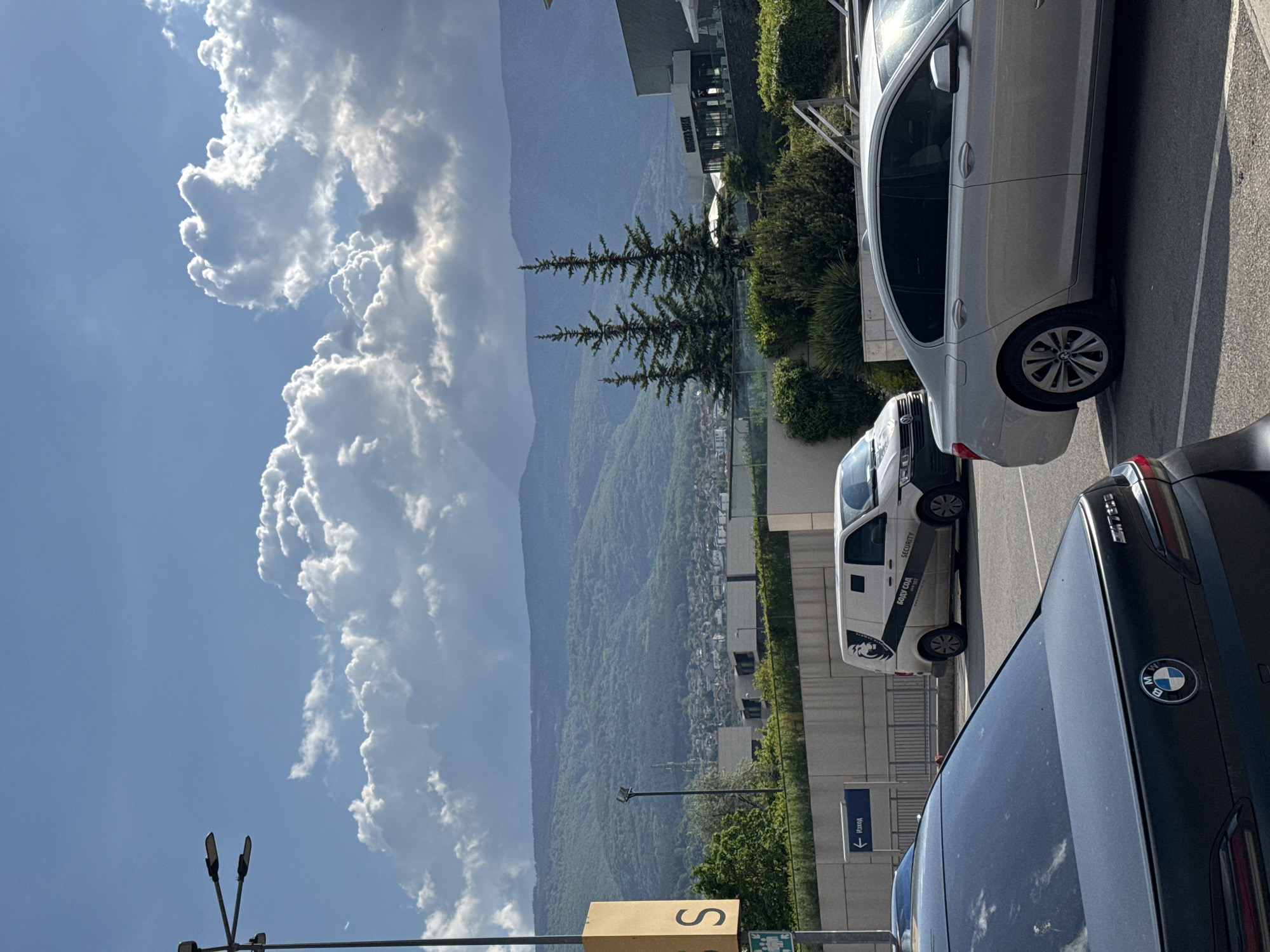 The image depicts a daytime scene in Sofia, Bulgaria. The view includes a mountain range in the background, partially obscured by a large cloud formation. In the foreground, there is a parking area with several cars: a black BMW, a white van marked with "Security" and "SODIV COA" in Cyrillic, and a silver sedan. A sign with the letter "S" and another sign with an arrow and "Изход" (Exit in Cyrillic) are visible. Streetlights are also present. The environment includes trees and greenery interspersed with urban buildings.