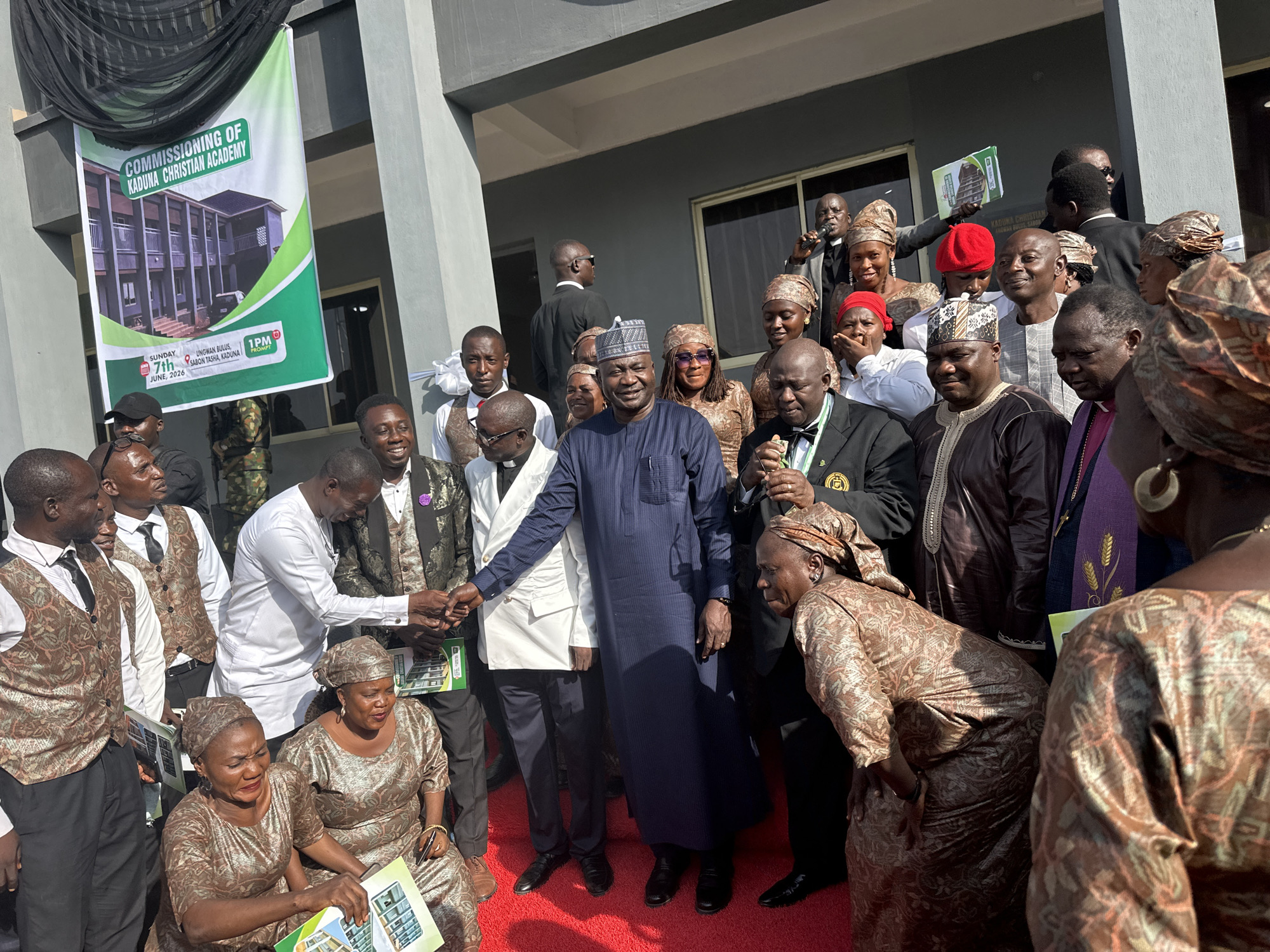 A large gathering of approximately 30-40 adults is depicted outdoors at the commissioning ceremony for the Kaduna Christian Academy in Ungwan Bulus, Sabon Tasha, Kaduna, Nigeria, as indicated by a prominent banner. The banner, colored green, white, and black, displays an architectural rendering of the academy building, the event title "COMMISSIONING OF KADUNA CHRISTIAN ACADEMY," the date "SUNDAY 7TH JUNE, 2026," the time "1PM PROMPT," and the specific location.
The ground where the people are assembled is covered with a vibrant red carpet. In the background stands a multi-story building with dark grey/blue exterior walls and numerous windows, presumably the academy itself or an adjacent structure. The scene is brightly lit by natural sunlight, casting discernible shadows.
The assembled individuals are primarily adults, with a diverse range of attire. Many men are wearing patterned brown and gold vests over white shirts and dark trousers. Several women are dressed in matching brown and gold patterned head wraps and dresses. A central figure, a man in a long dark blue traditional garment (agbada) and a patterned cap, stands prominently in the middle-ground. To his left, another man in a white long-sleeved shirt and dark trousers is engaged in a handshake with an individual largely obscured from view. A man in a black suit with a bow tie and a medal is visible to the right of the central figure. Further to the right, a man wearing a purple stole with wheat sheaf embroidery, indicative of clergy, is present. One woman in the mid-ground is wearing a red beret.
Many attendees are holding small, open brochures or booklets featuring an image of the academy building, mirroring the one on the main banner. On the far right, a man is holding a microphone, suggesting he may be speaking or officiating. In the foreground, two women in matching patterned dresses are seated or kneeling on the red carpet, also holding brochures. The participants appear engaged, with many smiling and looking towards the central activity.