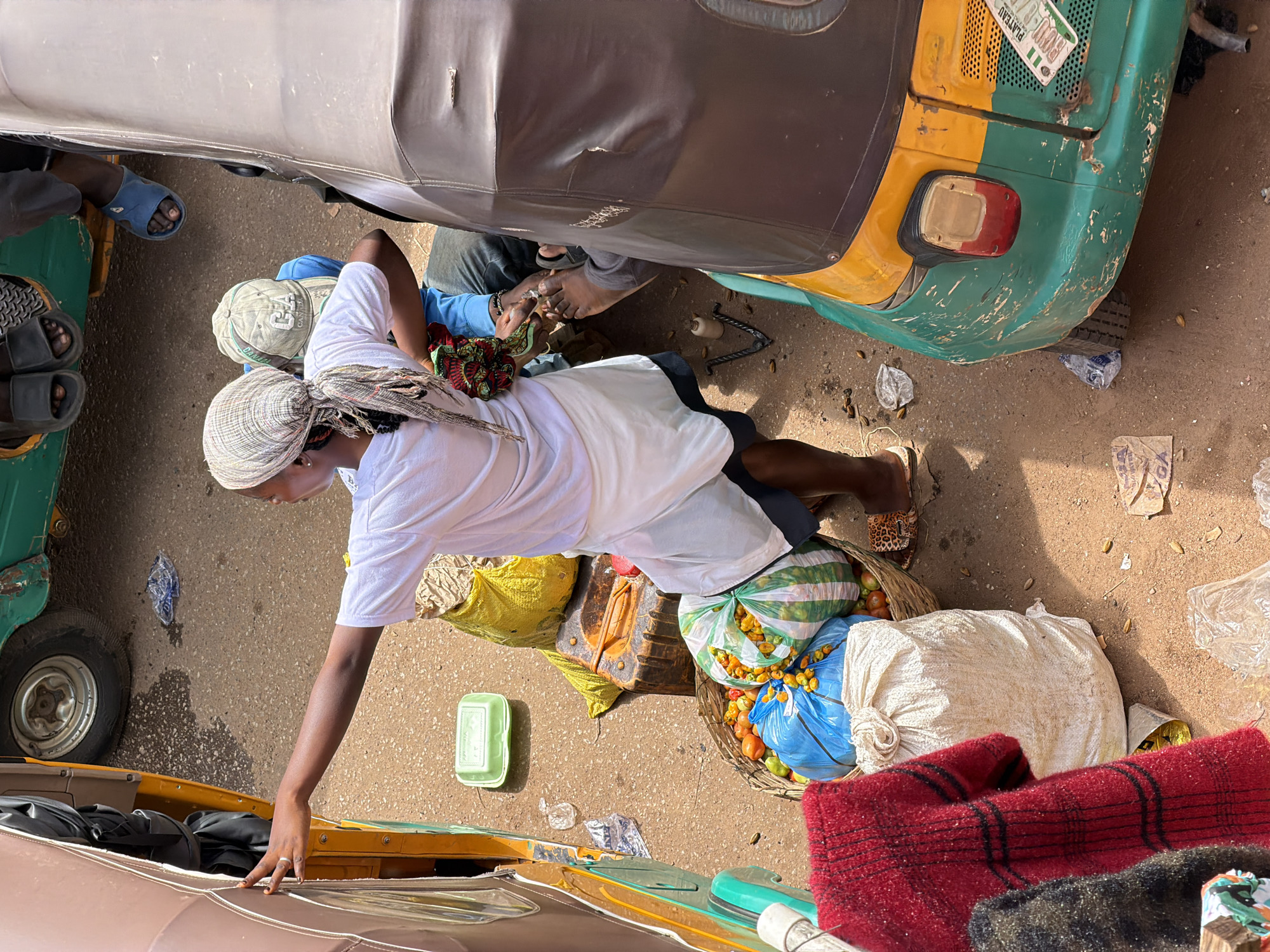 In Bukuru, Nigeria, a young woman, dressed in a white t-shirt and a light-colored headscarf, stands by the side of a beige and yellow auto-rickshaw. She is reaching out with her left hand towards the interior of the vehicle. In front of the auto-rickshaw, a basket overflows with tomatoes and small, yellow peppers. Beside the basket, a large white sack and a red and black checkered garment are visible. A green plastic container rests on the ground near the auto-rickshaw's front. In the background, a green auto-rickshaw is parked, with people's feet visible wearing sandals. The ground is a mixture of sand and dirt, with scattered debris.