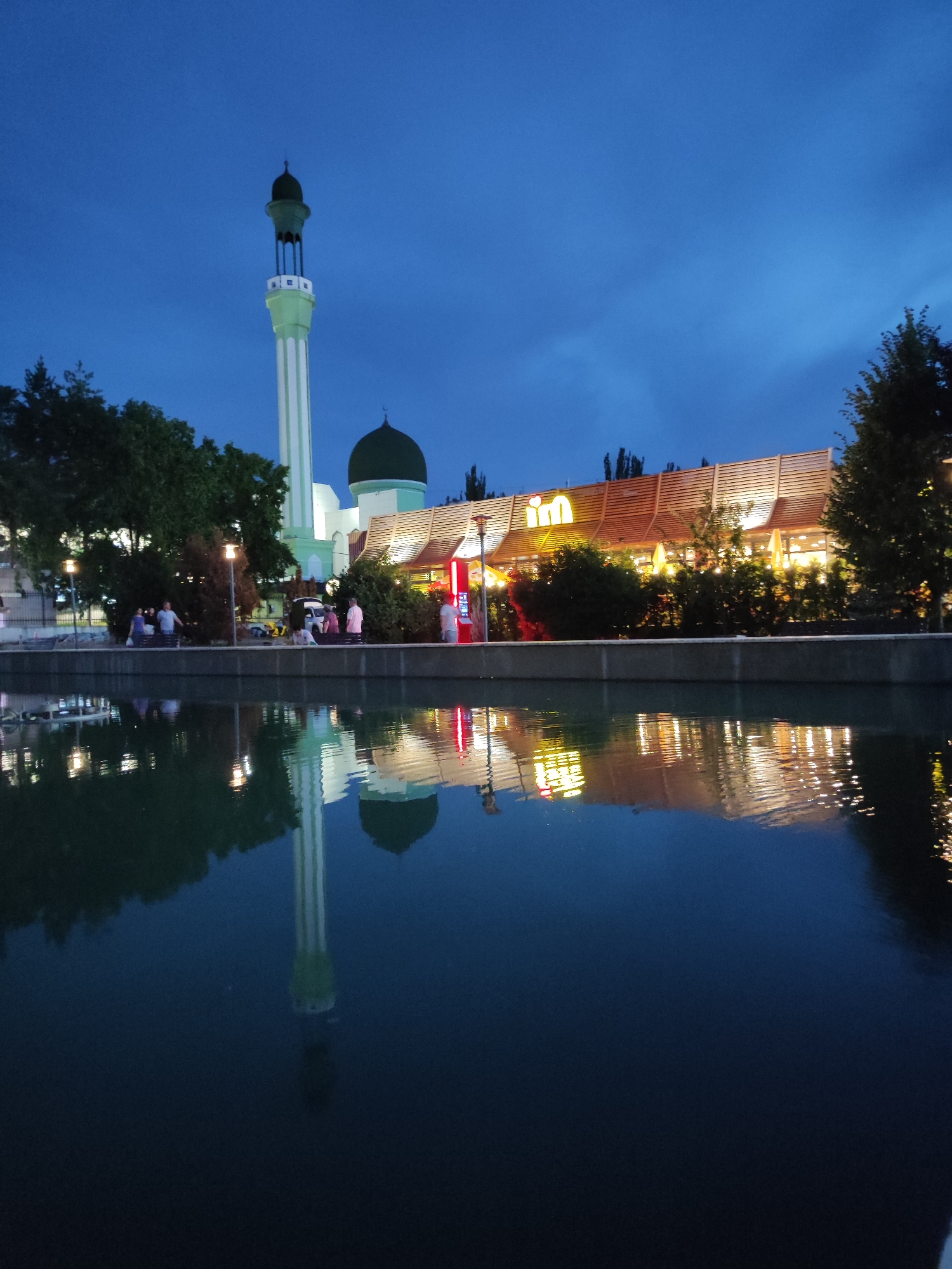 The image depicts a twilight scene in Almaty, Kazakhstan, with a mosque and a modern building reflected in a body of water. A tall, slender minaret, painted white with green accents, rises on the left side of the frame, topped by a green dome. Next to it, a larger green dome caps another section of the mosque. To the right of the mosque, a contemporary building with a wooden slatted facade is illuminated, featuring bright lights. The text "im" is prominently displayed in large, yellow illuminated letters on this building, likely indicating a commercial establishment.
In the foreground, the dark water of a canal or river perfectly mirrors the mosque, the minaret, and the illuminated building, creating a symmetrical and serene reflection. Along the embankment behind the water, a paved walkway is visible, with several people strolling or sitting on benches. Trees with dense foliage are interspersed along the walkway, some lit by streetlights. The sky is a deep blue, characteristic of dusk, with hints of lighter clouds. The overall mood is tranquil and peaceful, suggesting a calm evening in a public space. The presence of both religious and modern architectural elements, along with the reflective water, creates an interesting juxtaposition.