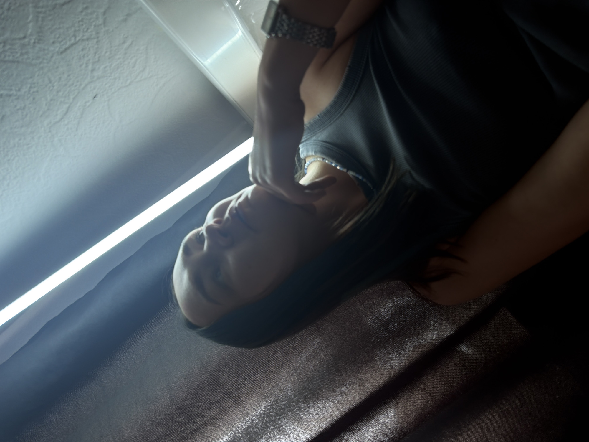 The image depicts a person indoors, likely in Chaikovsky, Russia, though this cannot be definitively confirmed from the image alone. The person is wearing a gray tank top and a metal watch. Their hair is dark. They are touching their face with their hand. There's a fluorescent light fixture overhead and a textured wall or surface. The overall lighting is dim. There's a reflective dark-colored surface or fabric in the background.