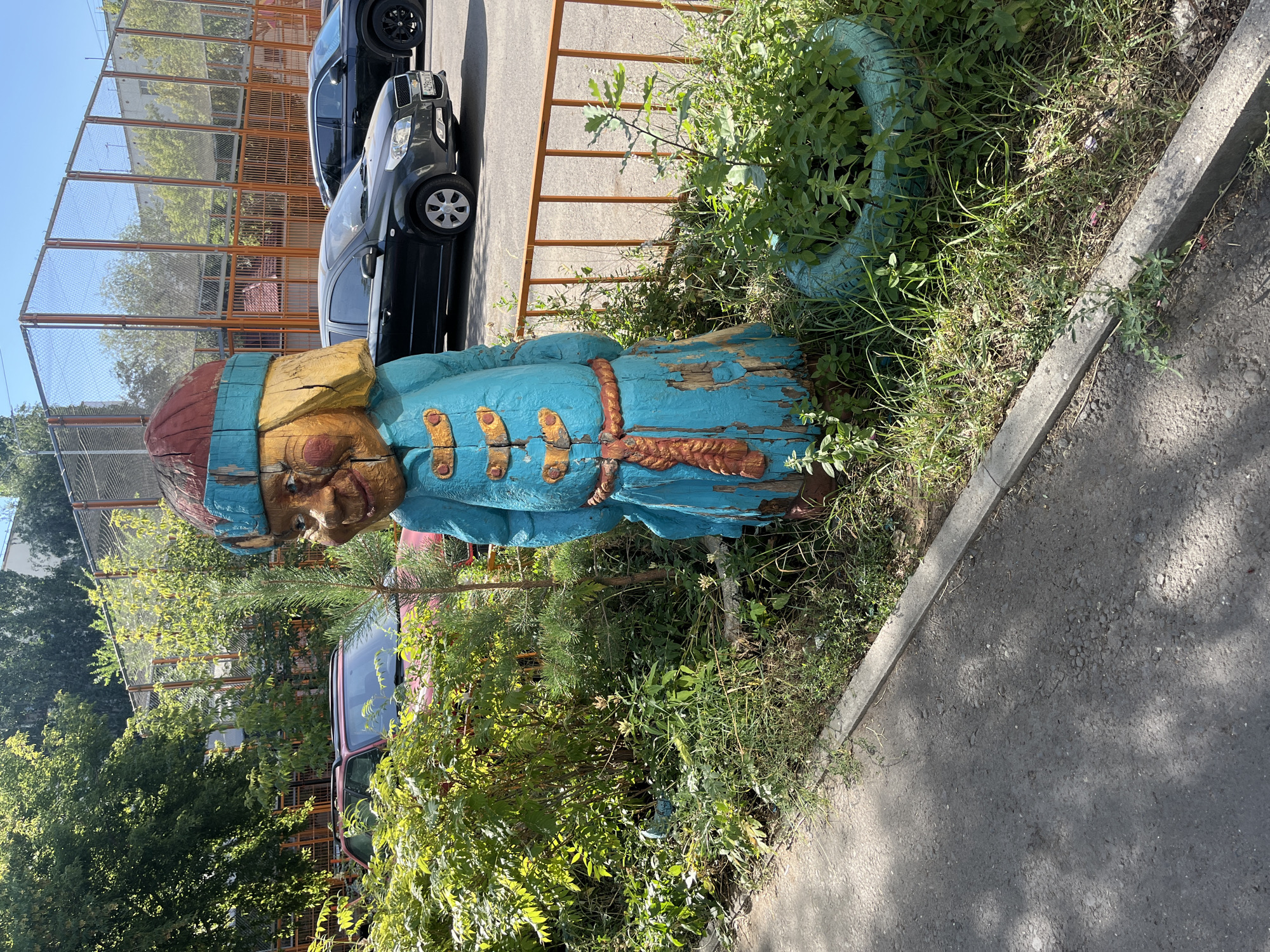 The image depicts a weathered, life-sized wooden sculpture of a figure wearing a blue tunic and a red and blue hat, positioned outdoors in a sunny environment. The sculpture exhibits signs of age and wear, with peeling paint and visible wood grain. It is surrounded by overgrown greenery, including bushes and tall grass, and a repurposed blue tire filled with plants serves as a planter. In the background, several cars are parked on an asphalt surface, and a chain-link fence surrounds a sports court or similar area. A concrete curb and sidewalk are visible in the foreground. The scene is likely located in Astana, Kazakhstan, suggested by the presence of parked vehicles and the overall urban outdoor setting.