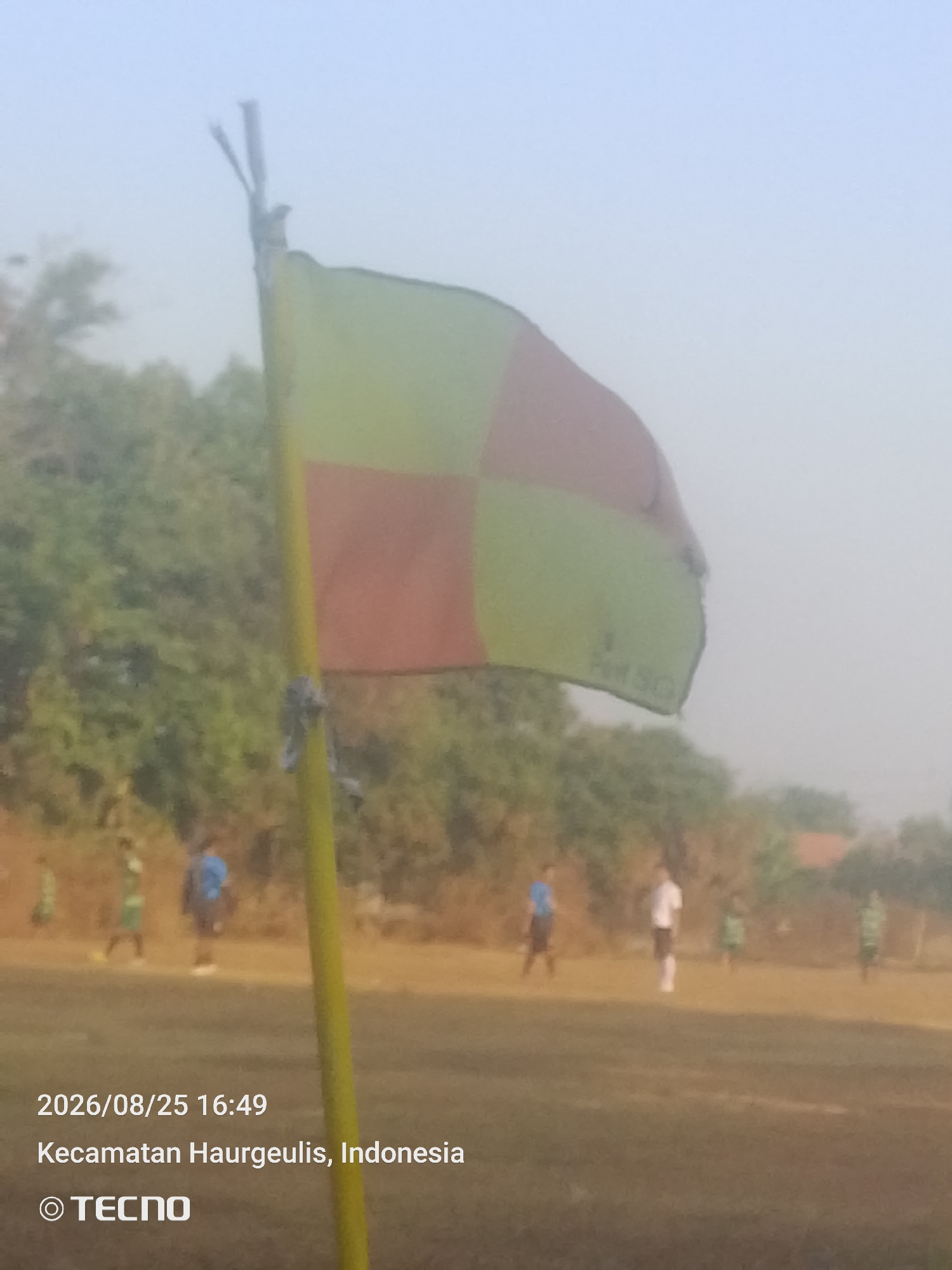The image captures a sporting event on a field in Kecamatan Haurgeulis, Indonesia, on August 25, 2026, at 16:49. In the foreground, a yellow flagstaff stands prominently, topped with a bicolor flag divided into red and yellow quadrants, which appears to be a corner flag from a football match. The flag is slightly frayed. In the background, several individuals are scattered across the field, some wearing athletic attire, suggesting a game or practice is underway. The environment is an outdoor field with a dusty or dry surface, bordered by trees and vegetation. The sky is a pale blue, indicating diffused light, possibly due to haze or early morning/late afternoon conditions. The TECNO logo is present in the bottom left corner.