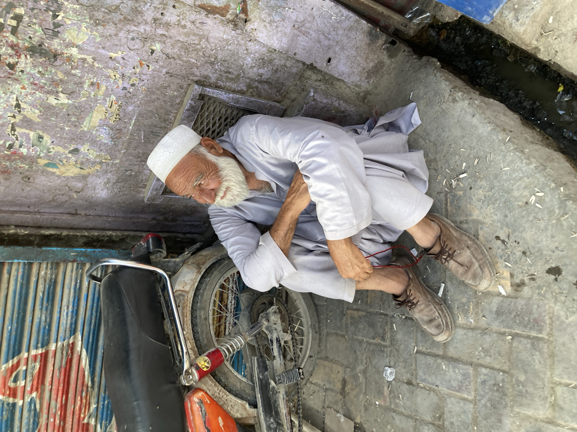 An elderly man, distinguished by a long white beard and a white prayer cap (koofi), sits on the ground in an outdoor setting in Quetta City Tehsil, Pakistan. He is dressed in a light-colored shalwar kameez and brown lace-up shoes. His face, etched with wrinkles, looks downwards, conveying a sense of pensiveness or weariness. His hands are near his feet, seemingly engaged with a red string or cord.
He is positioned beside a partially visible, well-used motorcycle with red and black components and a rusty rear wheel. The ground beneath him is a mix of irregular paving and concrete, heavily littered with debris, including what appear to be sunflower seed husks and cigarette butts. Behind him, a metal grate is set into the pavement, and further to the right, a narrow, dark drainage ditch is visible, containing murky water and refuse. The wall behind the man is weathered, with peeling paint and remnants of torn, faded posters. A fragment of text, "312 00", can be seen on one of these posters. The scene is brightly lit, indicating daytime, likely under clear weather. The overall environment suggests a bustling, yet aged, street corner, with an air of everyday life.