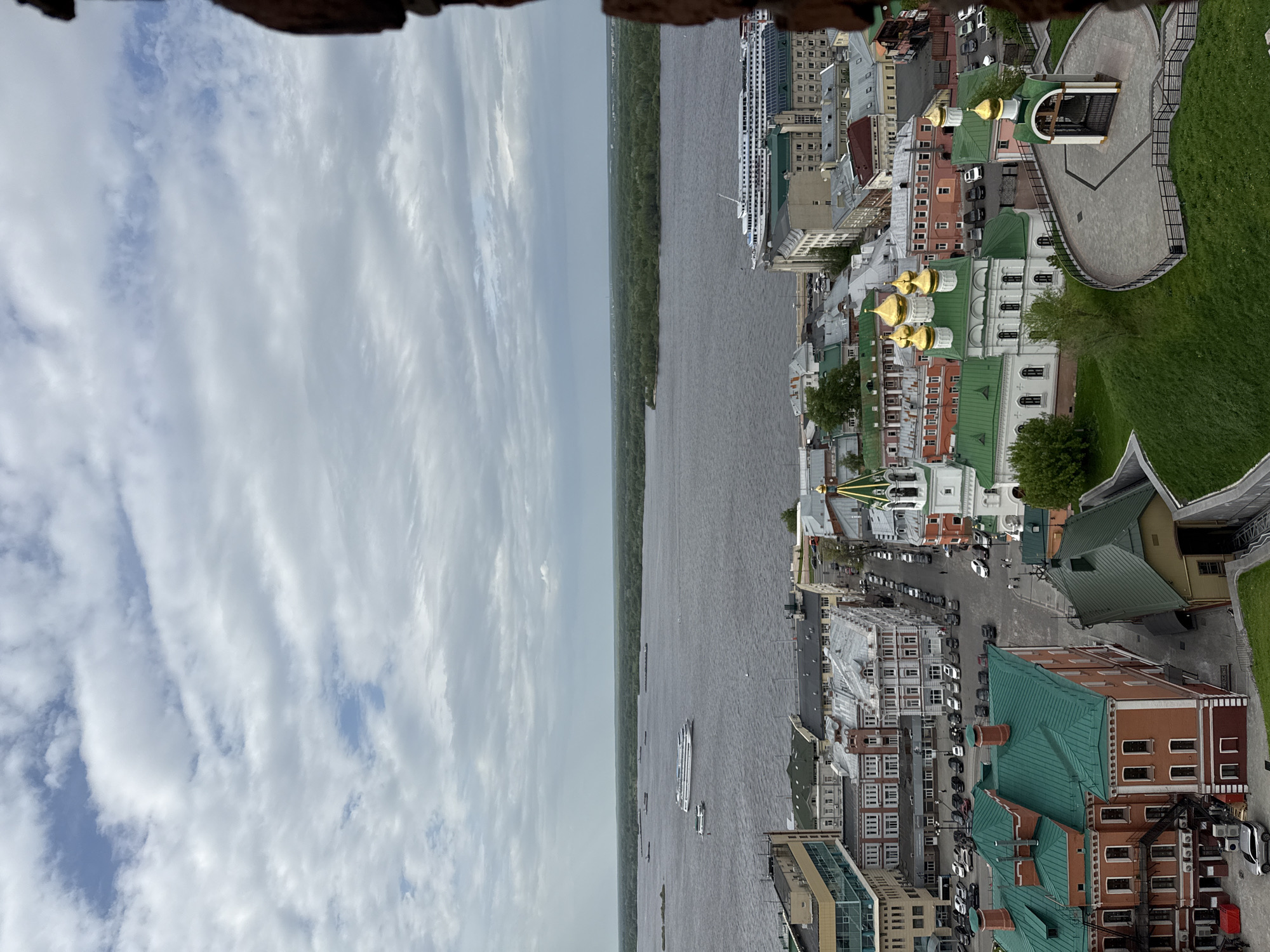 The image depicts an elevated perspective of Nizhny Novgorod, Russia, featuring the Volga River, various buildings, and architectural landmarks. On the left is the sky with scattered clouds. A wide river runs through the center of the frame, with a tree-lined shore on the opposite side. On the right, there is a view of the city with a mix of historical buildings, contemporary structures, and a glimpse of a green area. Buildings are varying in height and color, some with green roofs and golden domes. Cars and people can be seen along the city streets. In the river, there are several boats and possibly cruise ships. The image suggests a viewpoint from a high vantage point, such as a tower or building.
