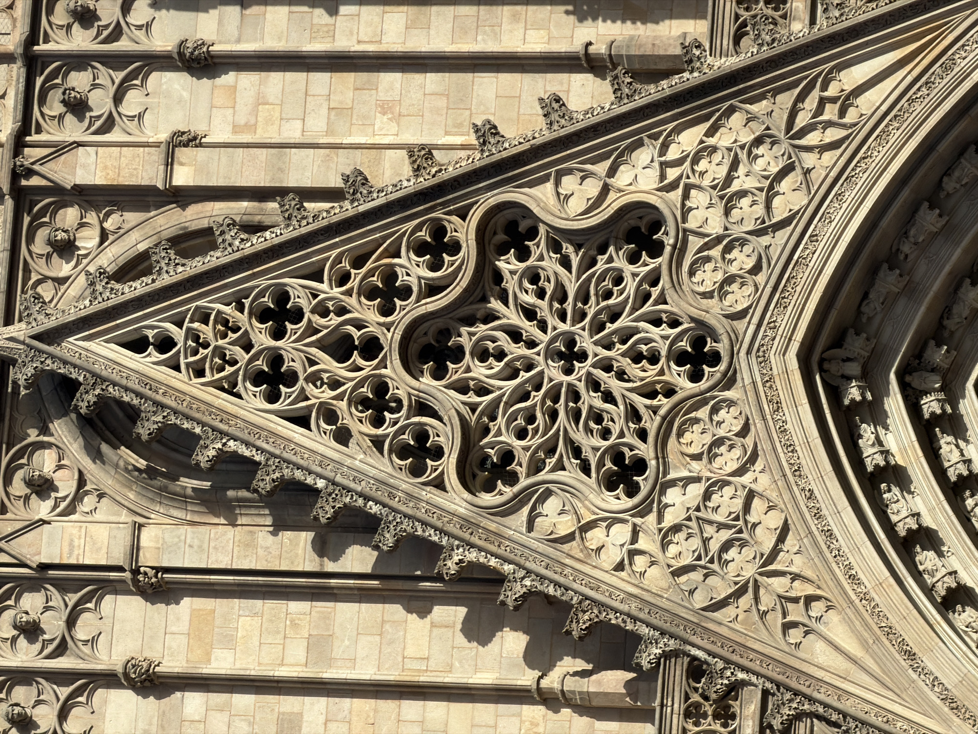 This is a detailed, close-up, eye-level, outdoor photograph of the ornate stone facade of the Barcelona Cathedral, located in the Gothic Quarter of Barcelona, Spain. The image focuses on a triangular section of the cathedral's exterior, featuring intricate Gothic architectural details.
The primary subject is a large, triangular stone carving with complex tracery work, reminiscent of a rose window or a gable. This carving is made of a light-colored stone, likely sandstone, and is deeply textured, creating strong contrasts with shadows. The tracery consists of multiple layers of stylized floral and geometric patterns, with repeated quatrefoils (four-lobed shapes) and curving lines forming a delicate lattice. The edges of this triangular section are adorned with a serrated, crenellated border, also carved from stone, with small, menacing gargoyle-like figures projecting outwards at intervals.
Behind the triangular carving, and framing it on the left, is a section of the cathedral's wall constructed from rectangular, light-colored stone blocks, arranged in courses. Vertical stone pilasters with decorative capitals break up the wall. Above and to the sides of the triangular carving, further architectural elements are visible. These include rounded arches, smaller decorative carvings, and what appear to be sculpted heads or busts enclosed within circular frames, set into niches above the main arch. These heads are carved in relief and wear simple, medieval-style headwear.
The lighting suggests it is daytime, with strong sunlight casting sharp, elongated shadows across the stone surfaces, highlighting the depth and complexity of the carvings. The overall impression is one of highly detailed, aged craftsmanship. No people are visible in the photograph. The environment is purely architectural, showcasing a section of a historic religious building.