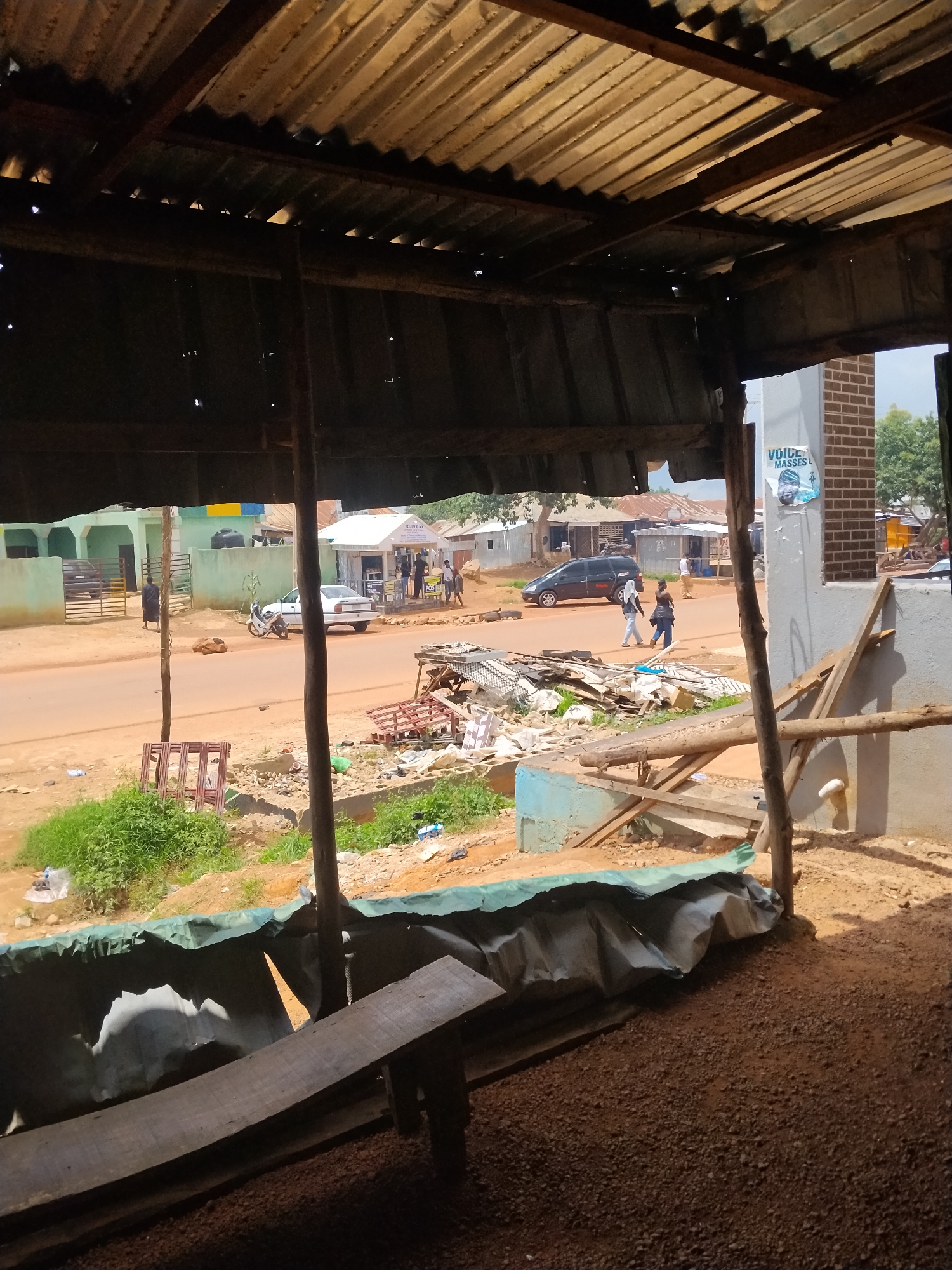 This is a street view in Durumi, Nigeria, taken from under a corrugated metal roof supported by wooden poles. The scene depicts a dusty, unpaved street lined with buildings. In the foreground, a rough wooden bench is visible. Across the street, there are several structures, including a light-colored building with a white awning and a brick building with a poster. A white car and a motorcycle are parked on the side of the road, and two individuals are walking on the sidewalk. The environment appears somewhat cluttered, with debris and building materials present. The overall impression is that of a modest urban or semi-urban setting.