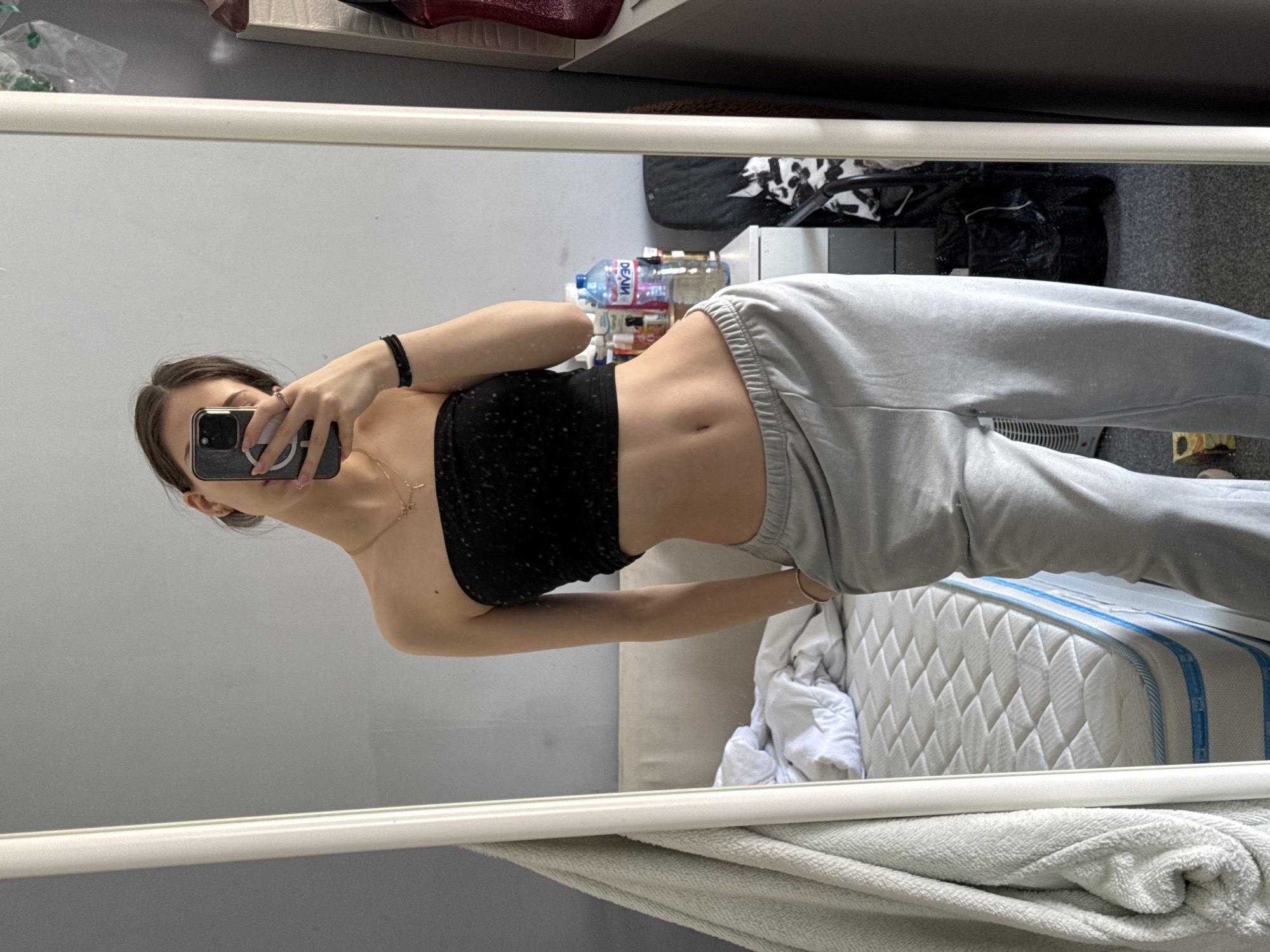 A young woman with long, dark hair is taking a mirror selfie. She is wearing a black tube top and grey sweatpants. Her torso is visible, showing her stomach. She is holding a smartphone in her left hand, obscuring part of her face. The reflection shows a cluttered room, including a bed with a white mattress, assorted items on a shelf, and a dark bag on the floor. There is no indication of Sofia, Bulgaria.