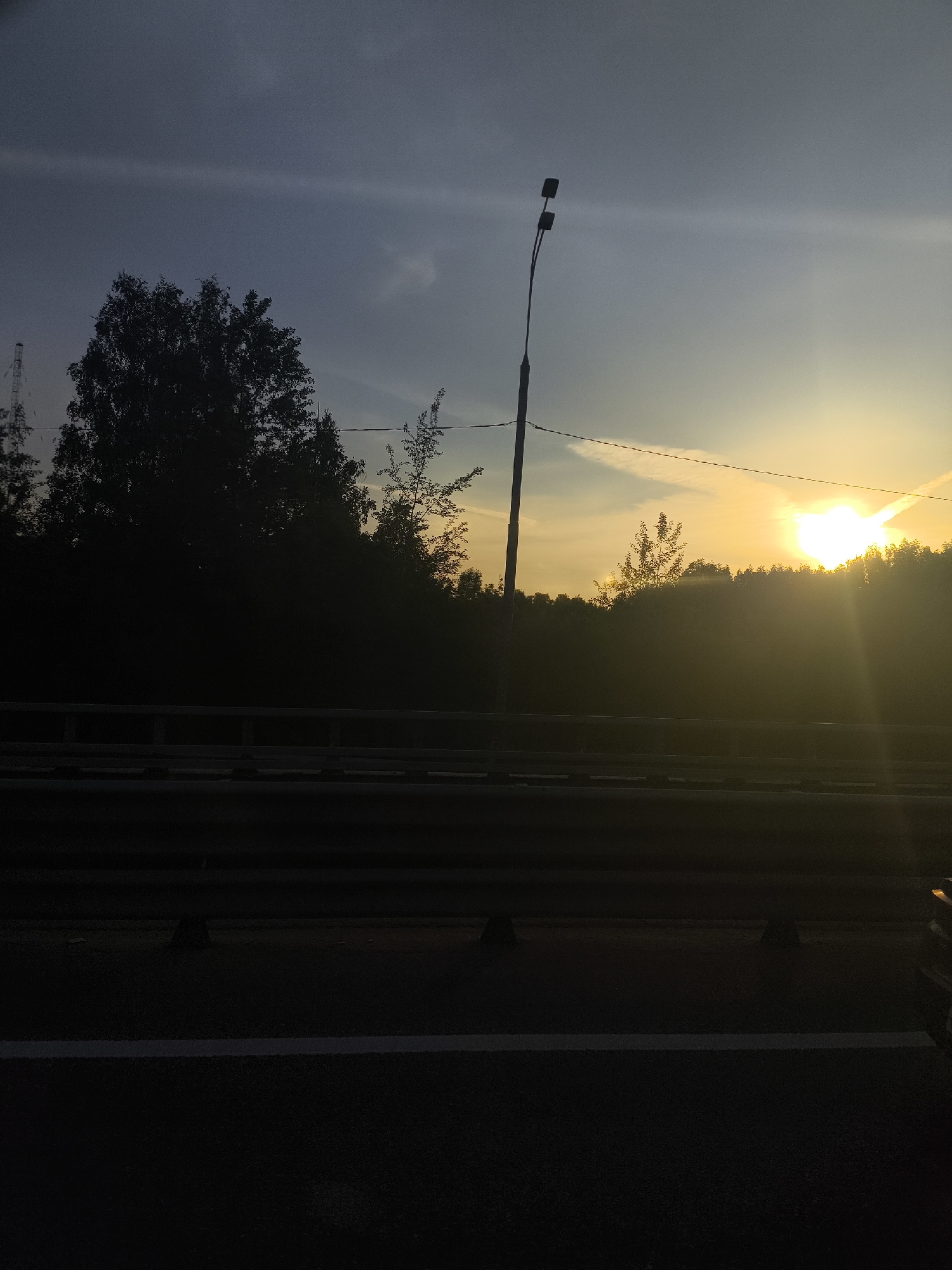 The image depicts a scene at dusk or dawn, potentially on a highway or elevated roadway. The foreground is dominated by the dark asphalt of the road with a white line marking its edge. A guardrail runs parallel to the road, obscuring some of the view beyond. Behind the guardrail, a dark silhouette of trees and foliage is visible against a sky that transitions from a pale blue at the top to a bright yellow-orange near the horizon where the sun appears to be rising or setting. A utility pole with two lamp fixtures stands tall among the trees. In the distance, an antenna or tower structure is barely discernible amidst the foliage. The location is Unknown, Unknown. No people are visible in the image.
