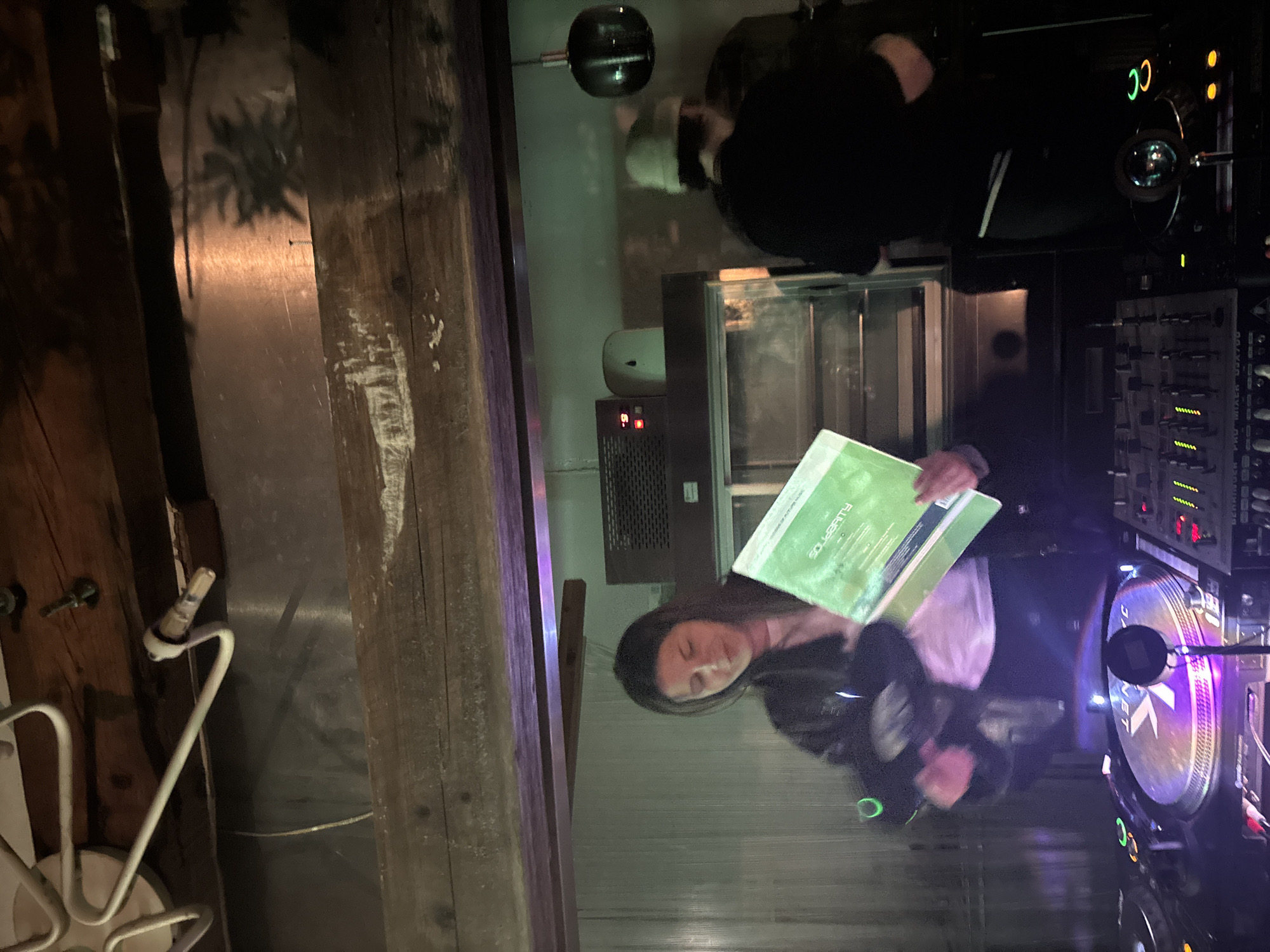 The image shows a person with long dark hair standing at a DJ setup, holding a green record sleeve with "SOLIDARITY" printed on it. The DJ setup includes a turntable with a purple illuminated disc and a mixing board with various dials and sliders. The lighting is dim, with some colorful accent lights. In the background, there's another person standing near what appears to be some equipment or shelving. The surroundings are somewhat cluttered, suggesting an industrial or warehouse space. Without further information, it is impossible to confirm the location in Lisbon.