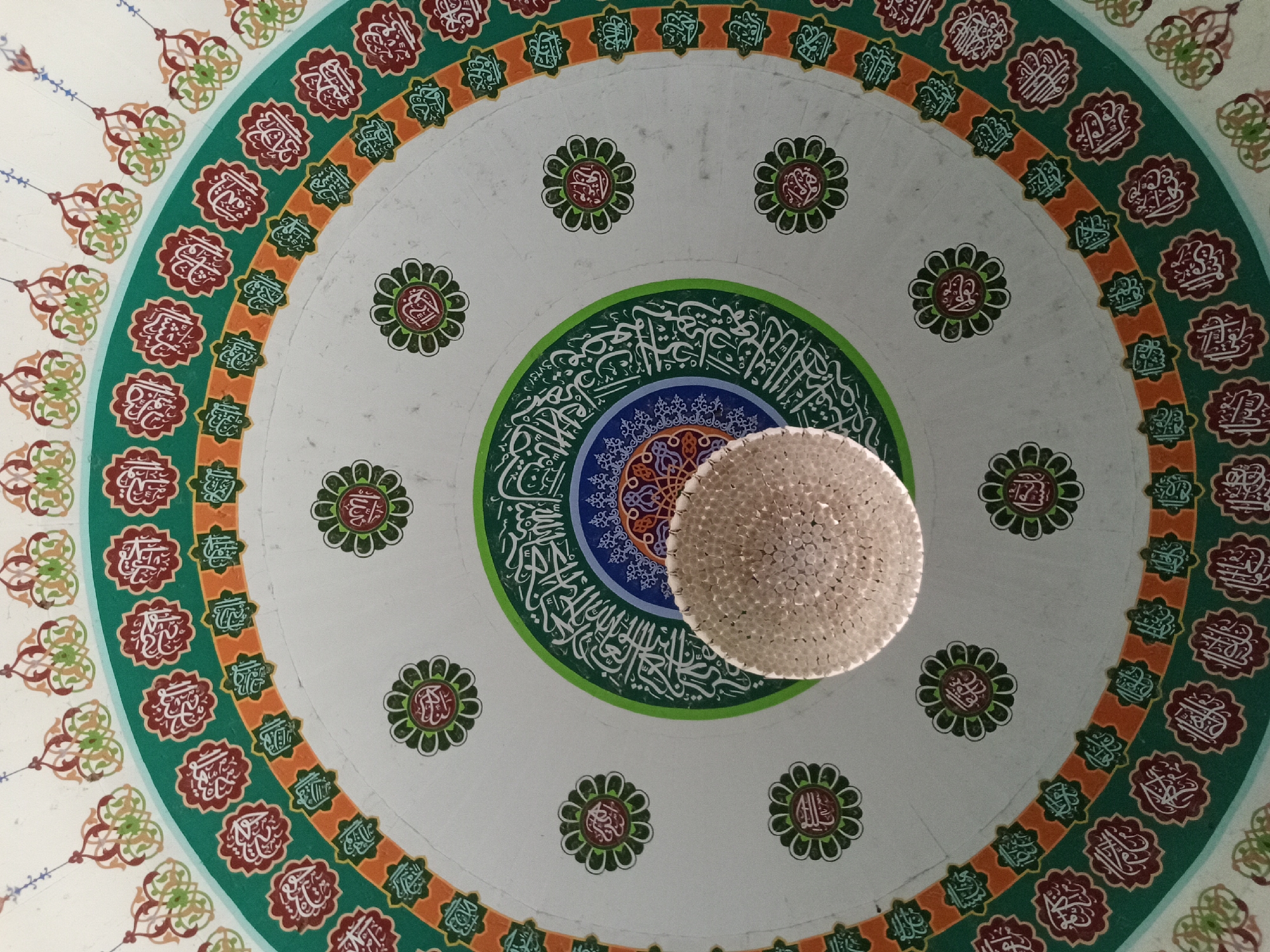 The image shows the interior of a building, likely a mosque, featuring a decorative ceiling. The ceiling has a circular design with concentric rings of patterns and Arabic calligraphy. At the center of the ceiling hangs a spherical chandelier. The location is Jantho, Indonesia.