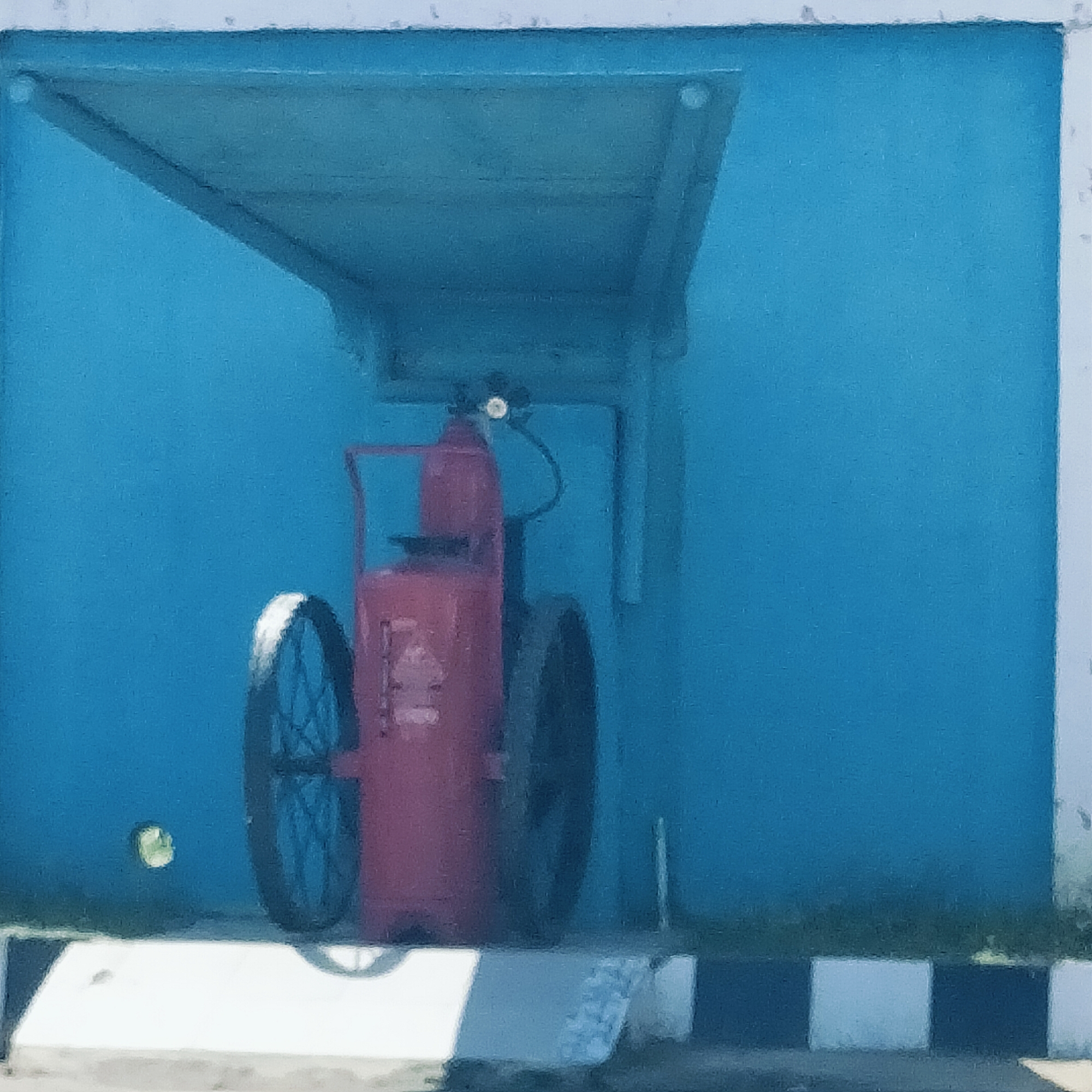 A large, industrial-sized, red wheeled fire extinguisher is the central subject of this image, positioned within a vibrant blue, open-fronted shelter. The extinguisher features two prominent spoked wheels, a top handle, and a pressure gauge, with a hose and nozzle assembly visible.
The shelter is a simple, shed-like structure with a flat roof and a back wall, all painted a striking blue. Small, circular reflections or fixtures are visible on the blue wall, one on the left and another on the right, beneath the roof. The blue wall is bordered by a white frame along its top and right edges.
In the foreground, a distinctive curb or barrier is visible, characterized by alternating black and white rectangular blocks. The ground inside the shelter and below the extinguisher appears to be concrete or dirt, partially in shadow. The scene is brightly illuminated, indicating daytime and clear weather. No people are present, and there is no discernible text on the fire extinguisher. The setup suggests a safety installation in an outdoor setting, possibly an industrial or public area in Cotpuuk, Indonesia.