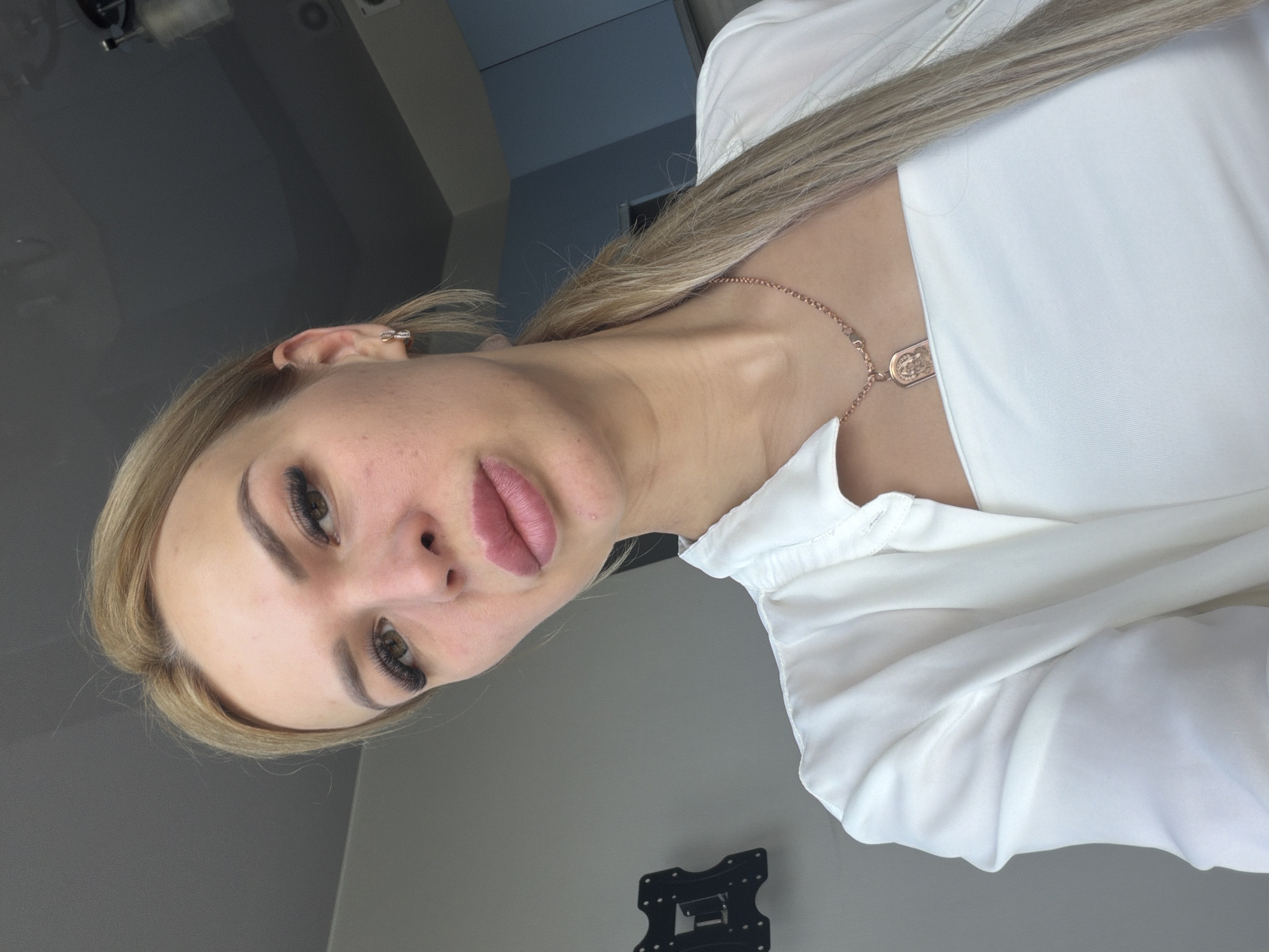 A young blonde woman is taking a selfie in an indoor setting. She is wearing a white top and an open white blouse. A rose gold necklace with a pendant is visible around her neck. Her hair is pulled back into a ponytail. In the background, a grey wall with a black TV mount is visible, as is part of a light blue cabinet. The ceiling is a neutral grey. The lighting suggests it is daytime. The scene is consistent with an apartment or office interior in Kyiv, Ukraine.