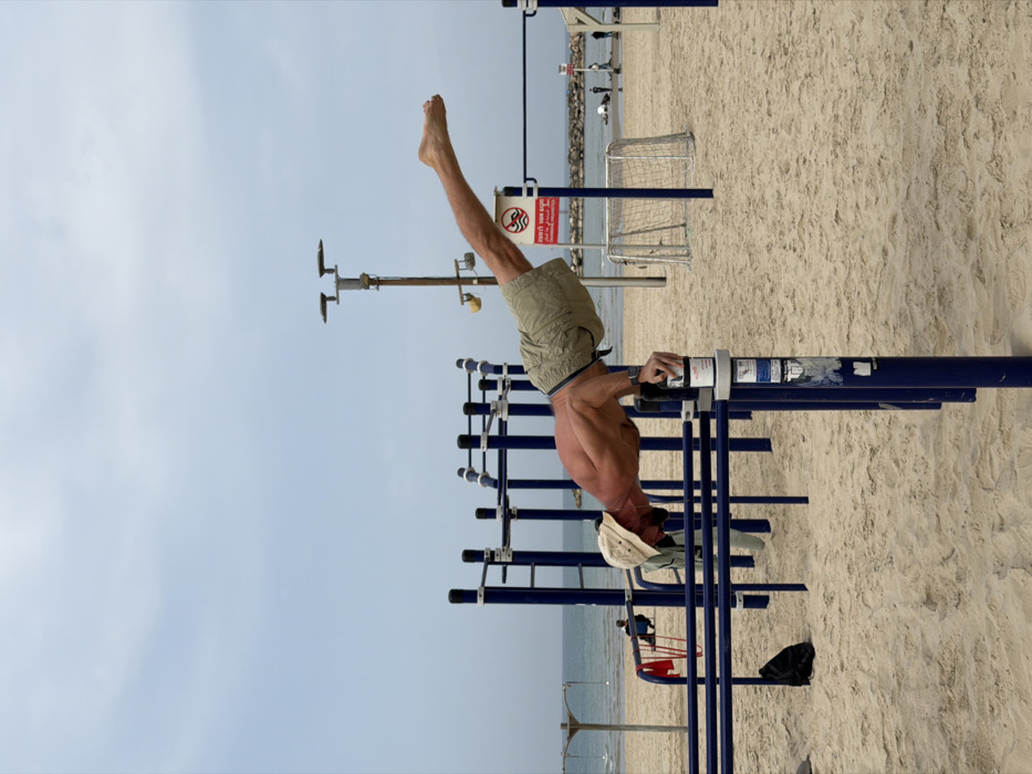 An outdoor calisthenics gym is depicted on a sandy beach in Tel-Aviv, Israel, under an overcast sky. The central figure is a shirtless, muscular adult male with a beard and a beige bucket hat, actively performing a horizontal plank-like exercise. He supports himself with his right hand gripping a white cylindrical container resting on a vertical blue bar of the calisthenics equipment. His body is extended straight, parallel to the ground, with bare legs stretched to his right. He wears light-colored cargo shorts and a dark wristband on his left arm.
The blue metal calisthenics structure occupies the foreground and mid-ground. To the left, a red resistance band hangs from a bar, and a black backpack rests on the sand. In the background, the Mediterranean Sea is visible, with a rocky breakwater in the distance. The sandy beach extends towards the water, populated by several indistinct human figures. A red circular sign with Hebrew text (indicating "No entry to water") is affixed to a pole to the right of the main subject, near a white portable soccer goal. A tall brown lamppost with two lights stands further back. The overall environment is a coastal exercise area.