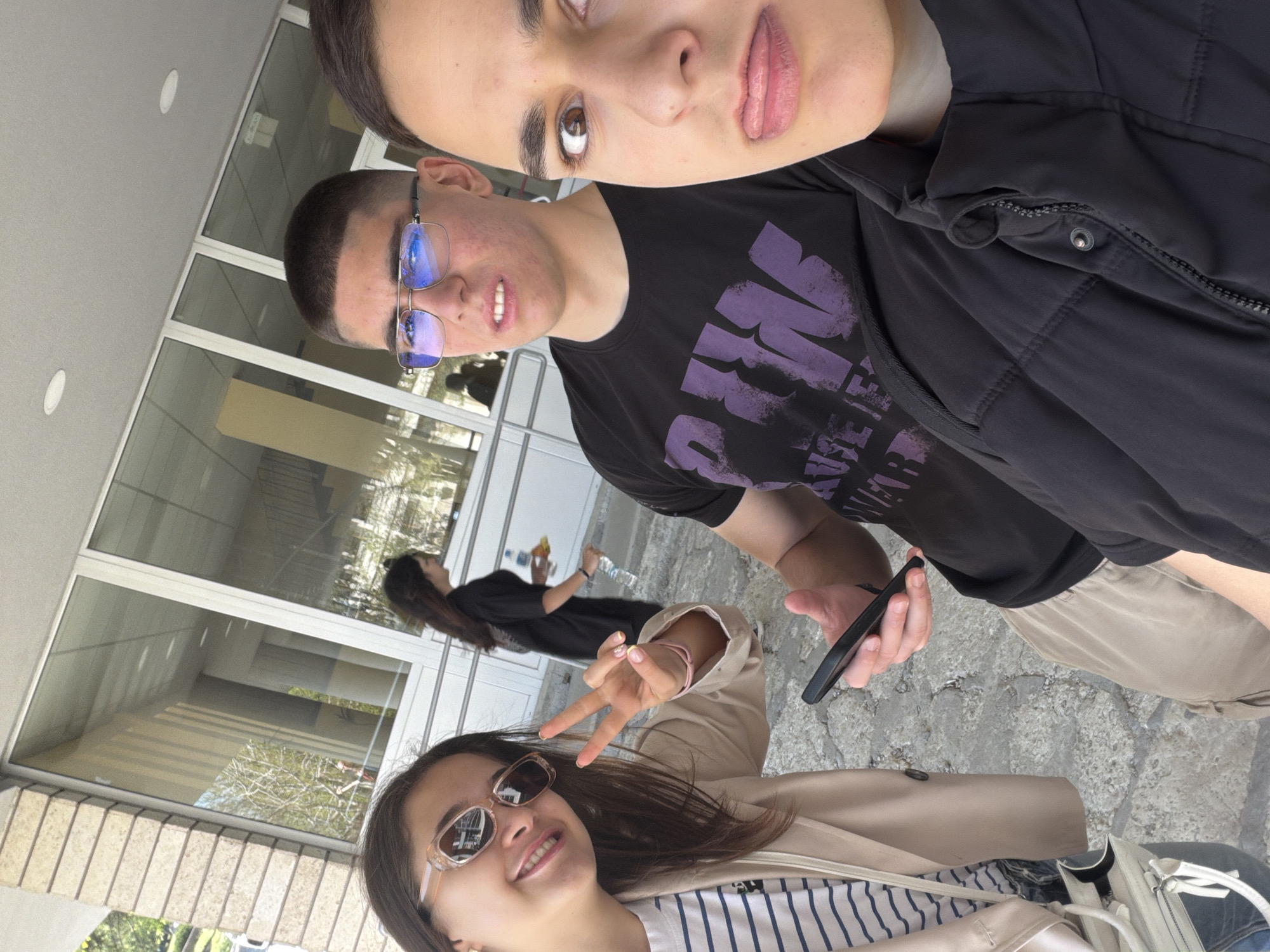 Three individuals are in an outdoor environment adjacent to a building. One person is wearing sunglasses and a striped shirt under a tan coat, displaying a peace sign. Another person is wearing glasses, a black t-shirt with purple writing, and holding a black phone. A third person is wearing a dark jacket. A fourth person, holding a water bottle, is further back and to the left. The background shows part of a building with windows and exterior stone or concrete paving. This location is NOT confirmed to be in the city of Shumen, Bulgaria.