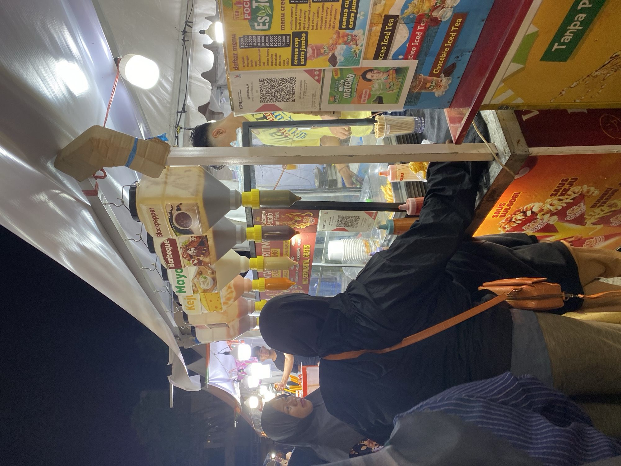 The image captures a nighttime scene at a food stall in Banjarmasin, Indonesia, likely a market or street food area. The stall is illuminated by artificial lights and features a row of dispensers for sauces and toppings, labeled with flavors like "Keju," "Maya," "Barbeque," and "Blackpepper." In the foreground, two individuals are visible, facing the stall, one wearing a black hijab and a black jacket with an orange shoulder bag, and another to their left wearing a grey hijab. Behind the counter, a young man in a bright yellow shirt is serving customers. Various menu boards and advertisements are displayed, including one for "Es Teh Poci" and "Choco Ice Tea." The overall environment suggests a lively, open-air food vendor setting.