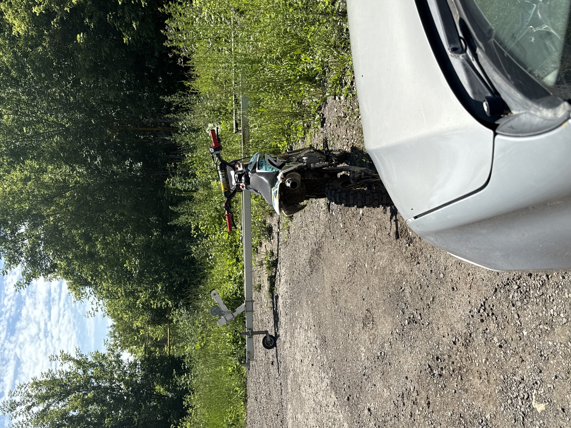 An outdoor shot depicts a dirt road with a dirt bike parked on it, facing away from the viewer. The bike is teal and black with red handlebars. A boat trailer is situated behind the dirt bike and to the left. The scene is set in a wooded area with lush green foliage and a bright blue sky with scattered clouds. The foreground shows the hood of a silver car. The location is identified as Samara, Russia. There are no people present.
