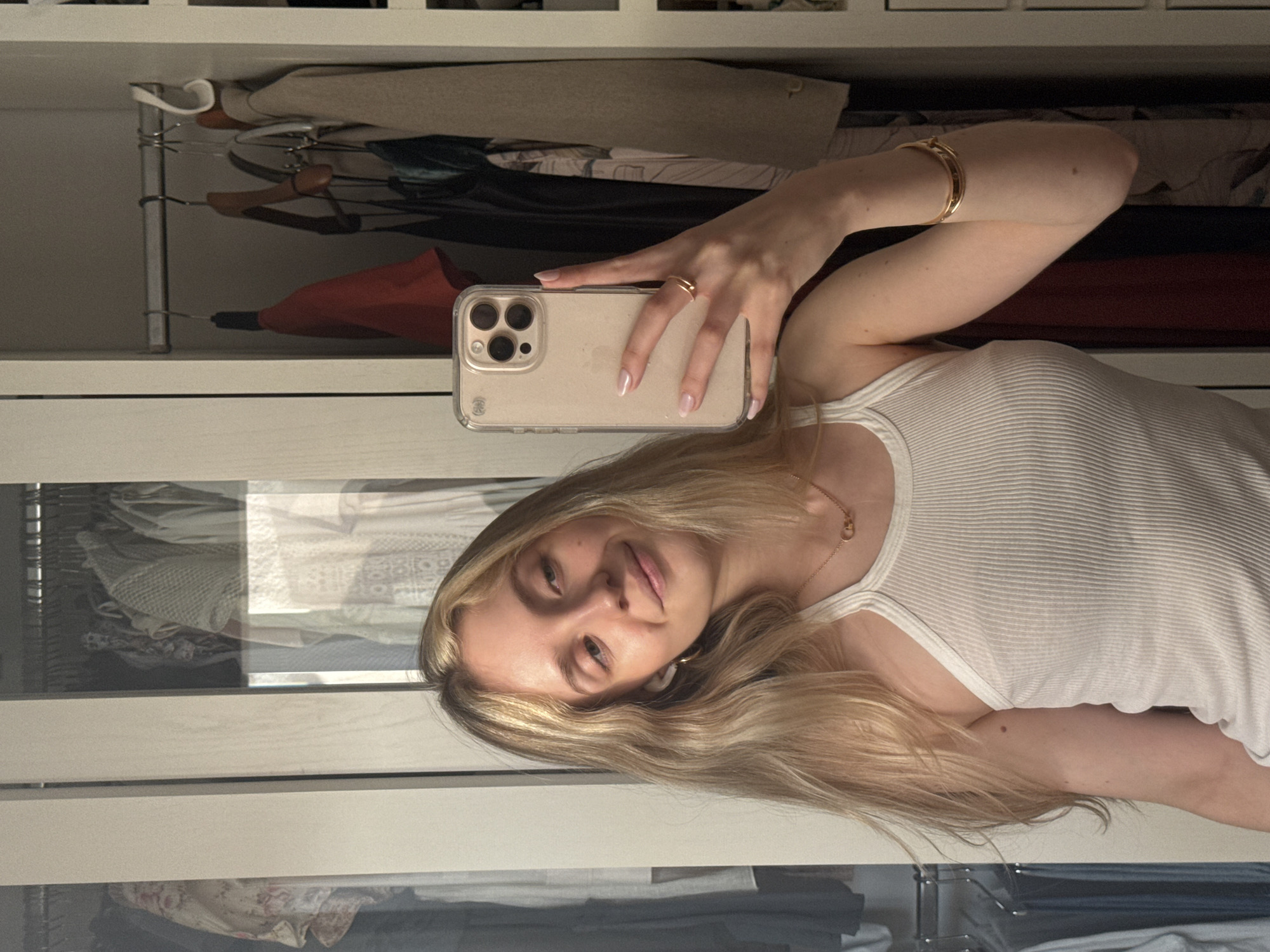 A young woman with blonde hair is taking a selfie in front of a closet. She is wearing a white tank top, a gold bracelet, and a ring. Her hair is styled and she has fair skin. She is holding a beige smartphone in her left hand. The closet is full of clothes, hanging on rods. The closet appears to have a white frame with a partially mirrored door. The location is not discernible from the image. The image does not reveal any information about being in Mexico City, Mexico.