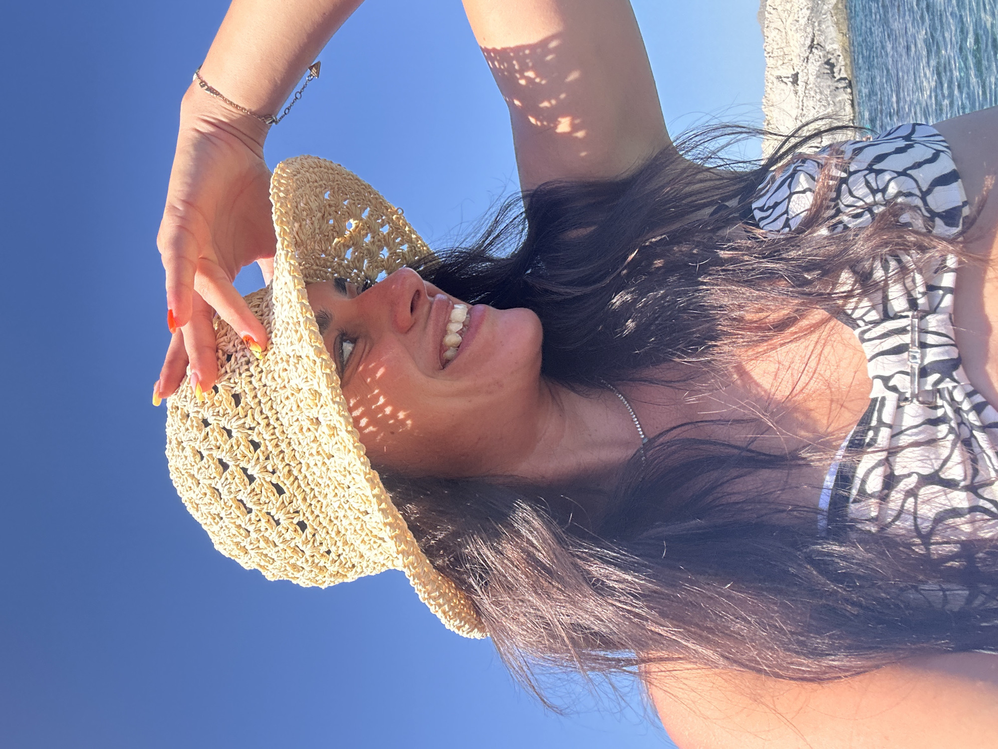 A woman wearing a woven straw hat and a black and white patterned bikini top is captured from the chest up, in a close-up shot. She is looking to her right, with her left hand raised to shade her eyes from the sun. Her long, dark hair is flowing around her shoulders. The background consists of a clear blue sky, the ocean, and a rocky coastline. The lighting suggests a sunny day, possibly in a Mediterranean location like Cala Lliteres, Spain, given the clear blue sky and the presence of rocky coastal features.