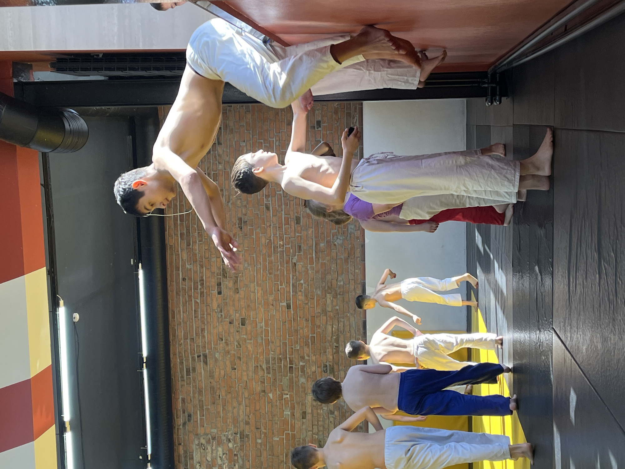 This image depicts an indoor training session in Krasnoyarsk, Russia, likely a martial arts or gymnastics class, with a focus on young male participants. The scene features approximately eight boys and teenagers engaged in various exercises.
In the foreground, two individuals are performing inverted maneuvers against a reddish-brown wall. One shirtless boy with dark hair is captured in a deep forward bend or handstand-like position, his body parallel to the floor. Above him, the lower legs of an adult, clad in white pants, are visible, suggesting they are spotting or instructing the boy. Another boy nearby is also in an inverted pose, wearing white pants.
Further into the background, a line of boys, mostly shirtless and wearing white or blue training pants, stand on a dark, possibly rubberized mat. They are positioned in a bent-over posture, facing a brick wall, indicative of a warm-up, stretching, or specific exercise routine. Bright sunlight streams into the space, creating strong highlights and shadows on the floor and walls.
The training environment features exposed brick walls, dark structural elements like beams and pipes, and fluorescent strip lighting. The overall atmosphere suggests a dedicated and focused training session during daylight hours. No visible text is present in the image.