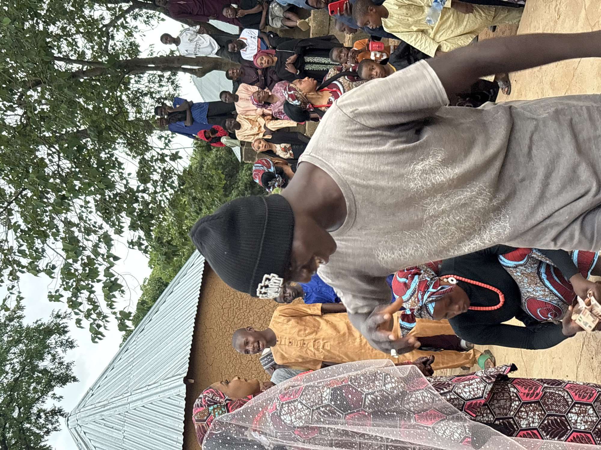 In Jos, Nigeria, on a cloudy day, a group of people gather outdoors. A young man in a grey t-shirt and black beanie is in the foreground, gesturing with his left hand. He is facing right, his mouth open as if speaking or laughing. To his left, a woman in a patterned headscarf and dress, with a veil over her head, is looking towards him. Behind them, a man in a light orange tunic stands and smiles, facing left. Further back, a crowd is gathered, many seated on tiered steps. They are dressed in various traditional African attire and Western clothing. The environment includes a mud-brick building with a corrugated metal roof, trees, and a cloudy sky. A woman in the middle ground, wearing a black top and a patterned headscarf and skirt, is kneeling and holding money in her hands.