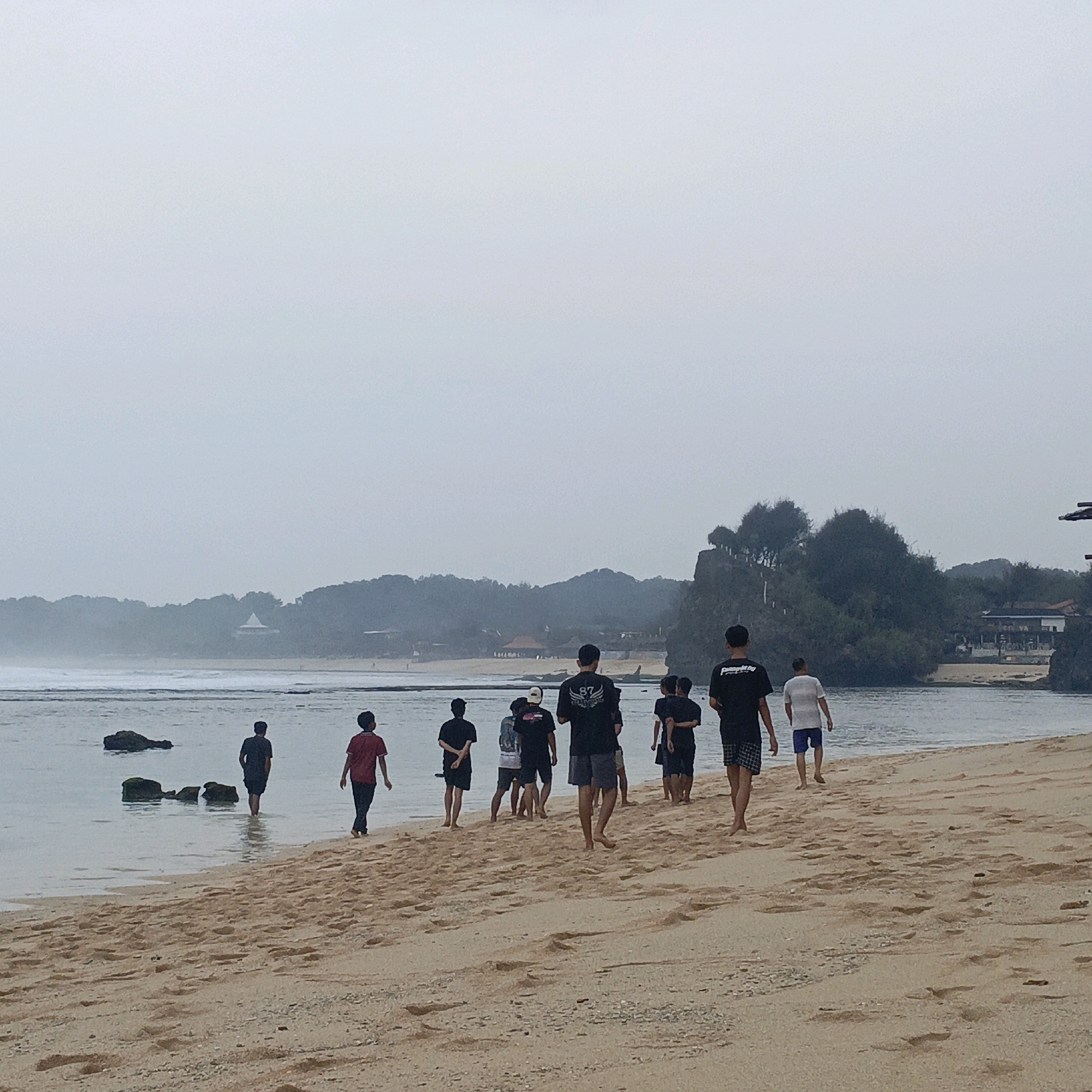 The image shows a group of young males walking on a sandy beach in Tepus, Indonesia, with the ocean and a tree-covered hill in the background. The sky is overcast. Several individuals are visible in the water, while others are walking along the shoreline. The individuals are dressed casually in t-shirts and shorts. The scene appears to be a leisure activity, possibly a walk or a gathering at the beach. The specific location within Tepus is likely a public beach accessible to visitors.